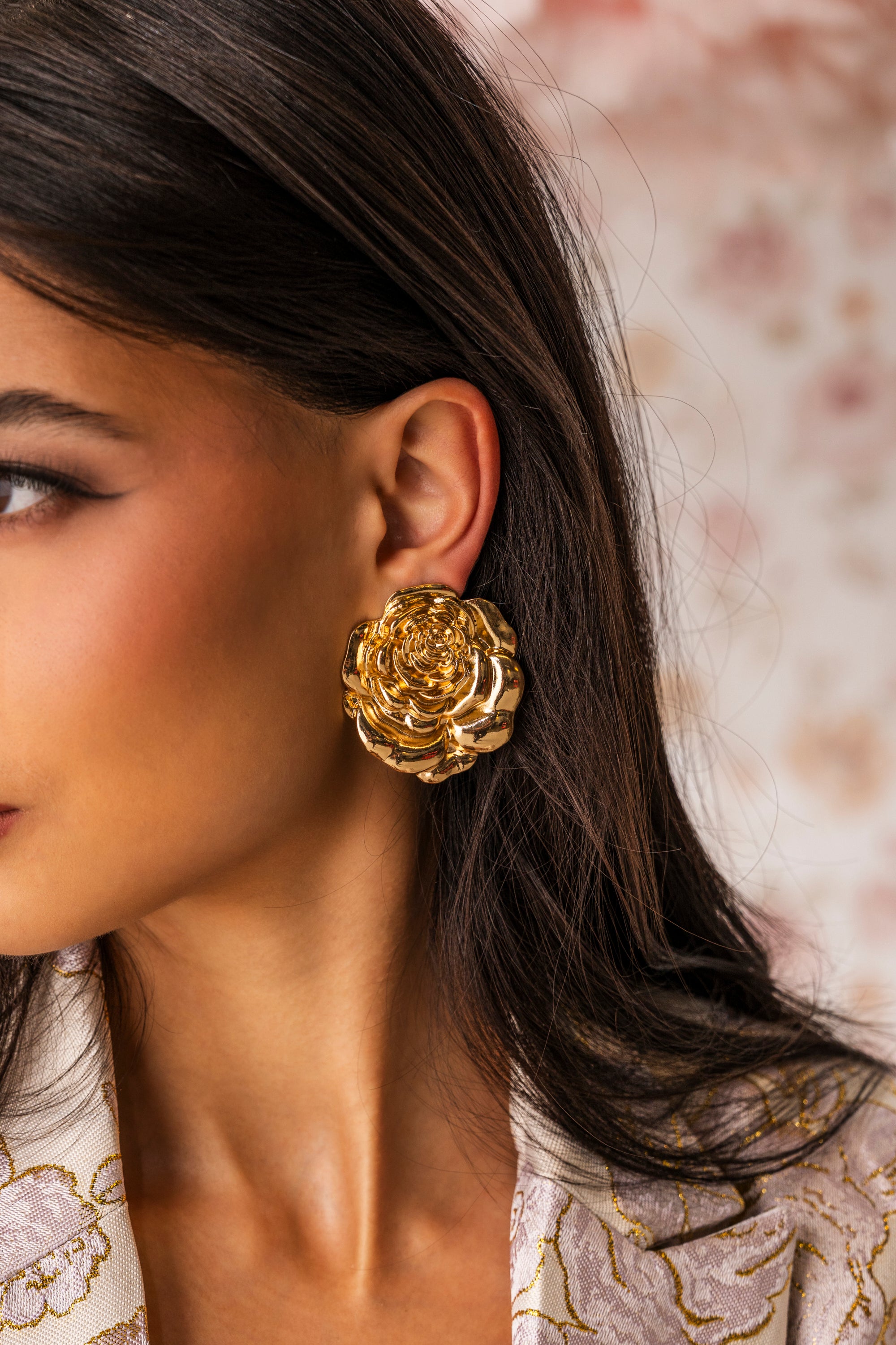 A close-up of a woman wearing IN BLOOM EARRING, large metallic gold floral earrings. She has long dark hair, smooth skin, and wears a cream garment with gold embroidery against an elegant, softly blurred background. 0