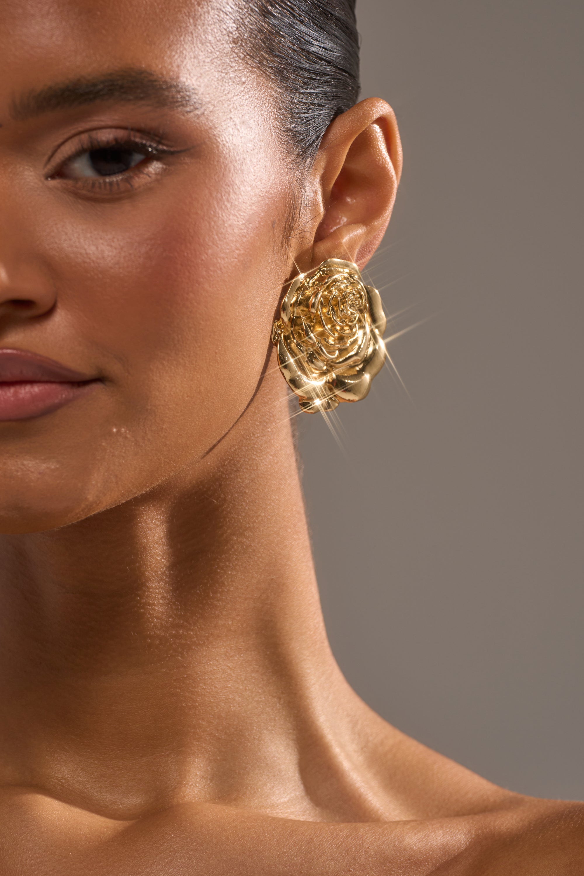 Close-up of a woman's face and neck showing the IN BLOOM EARRING—a shiny, gold metallic floral-shaped piece. Her smooth, glowing skin and slight smile stand out against a neutral, blurred background. 3