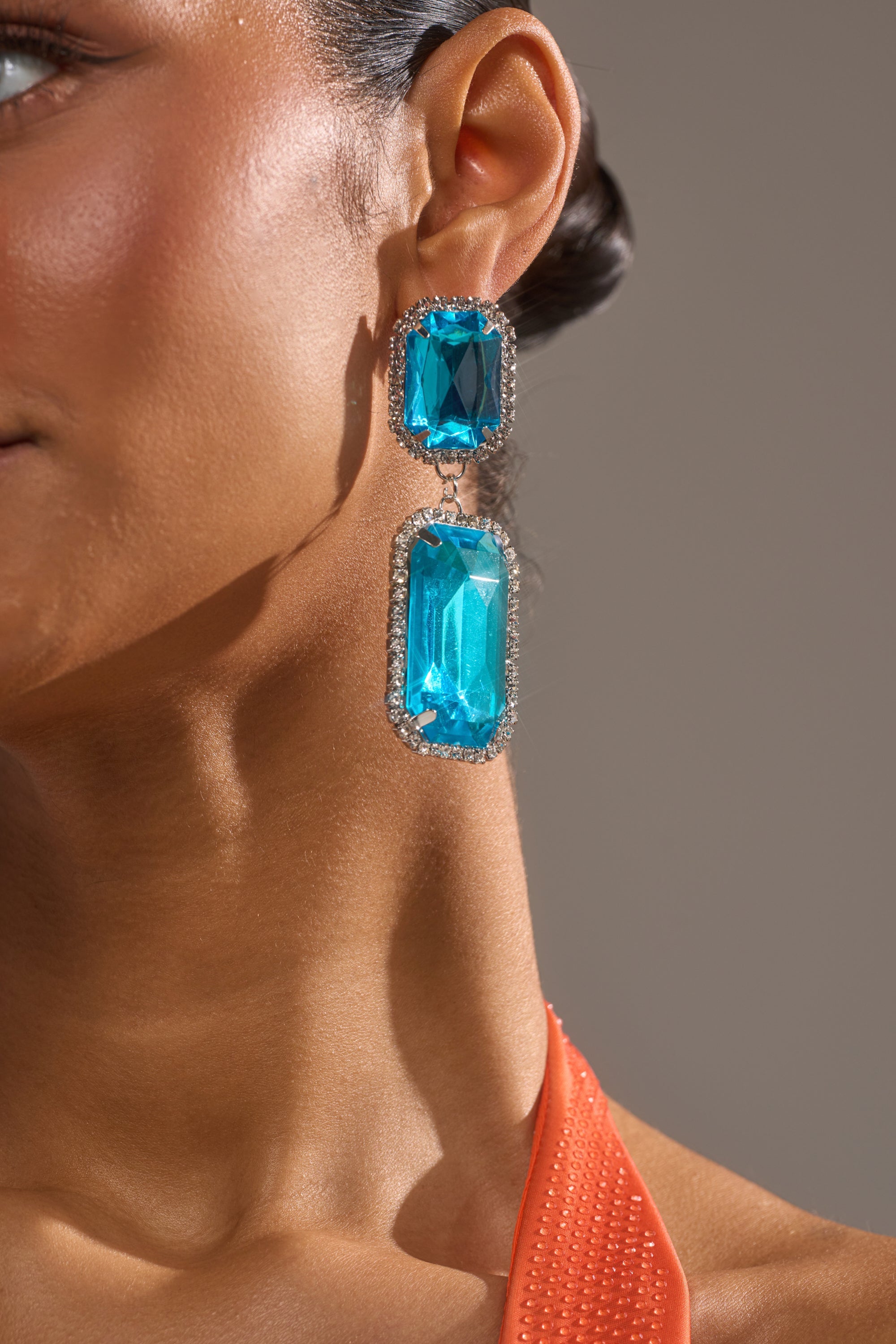 Close-up of a woman's ear wearing ON TREND EARRING, featuring two large rectangular turquoise stones bordered by rhinestones. She has smooth brown skin and an orange textured top. 2