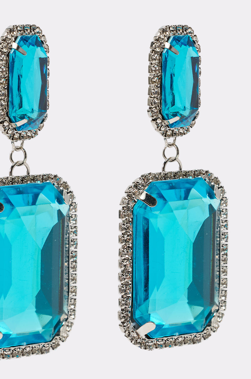 Close-up of the ON TREND EARRING: two silver dangle earrings with large rectangular turquoise gemstones, each framed by sparkling rhinestones for an elegant, glamorous look. 5
