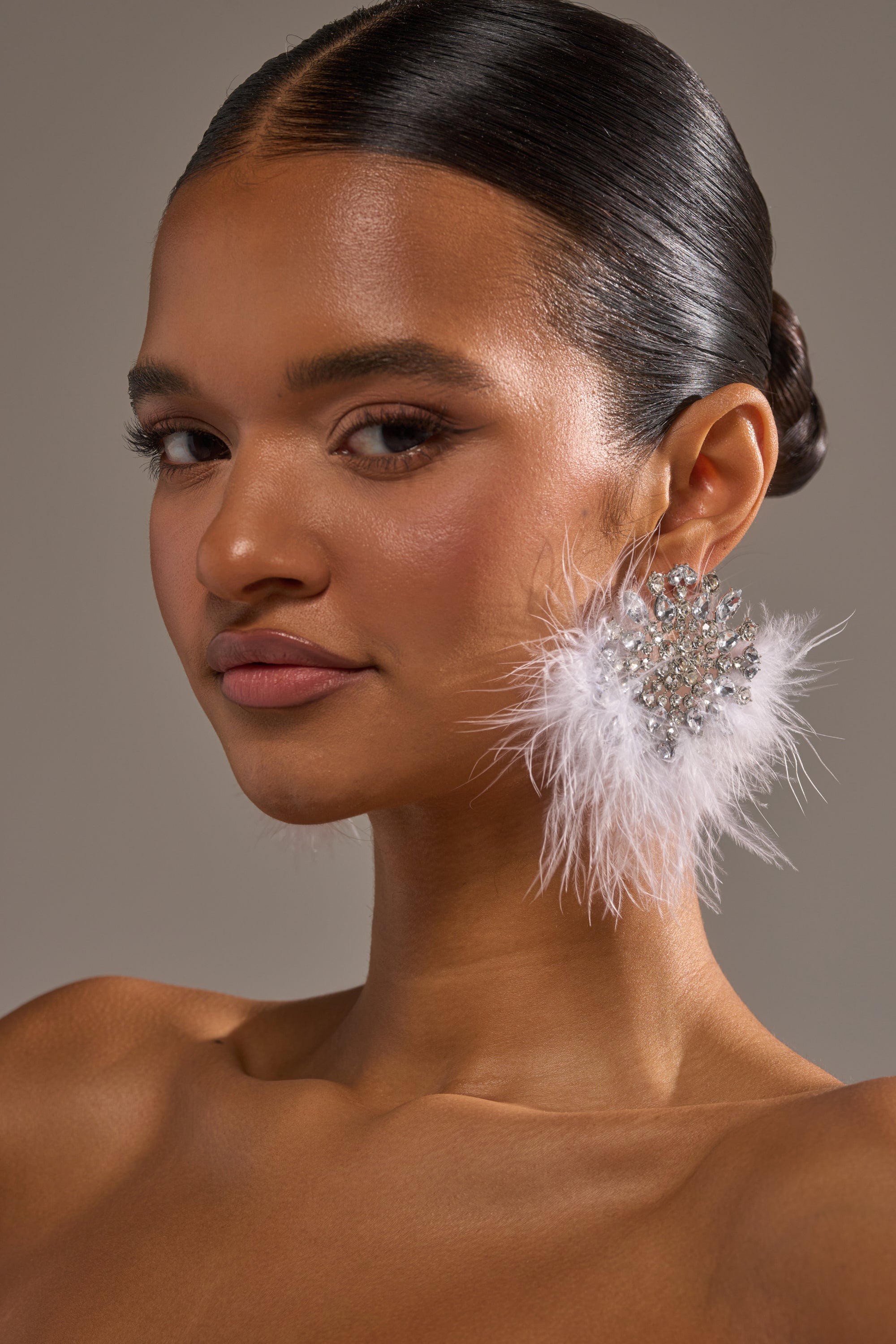 A woman with smooth, dark hair in a low bun wears the LIFE OF THE PARTY EARRING—sparkly, sunburst-shaped earrings with feather accents. Her glowing makeup and confident gaze stand out against a neutral background. 0