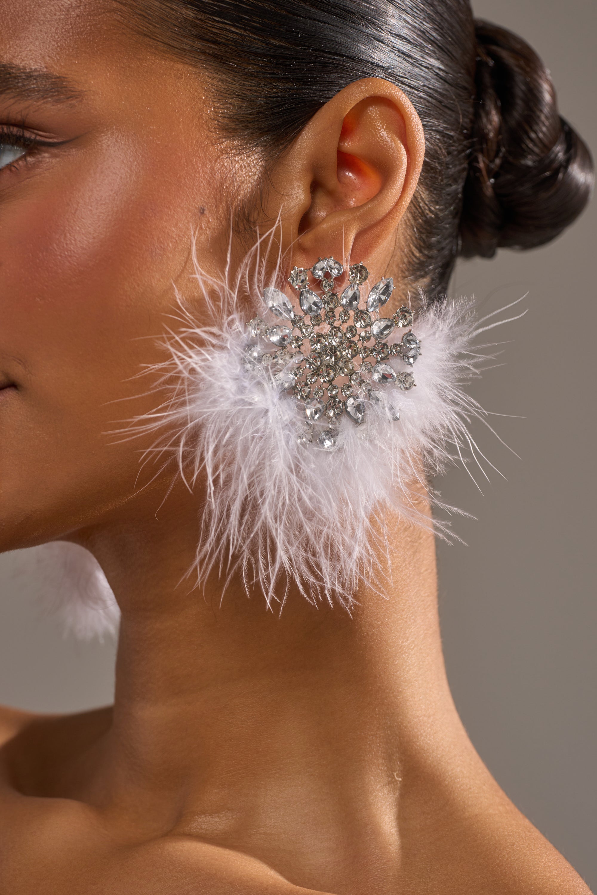 Close-up of a woman's ear wearing the LIFE OF THE PARTY EARRING, featuring sparkling crystals and white feathers. Her hair is in a sleek bun, her glowing skin set against a neutral background. 1
