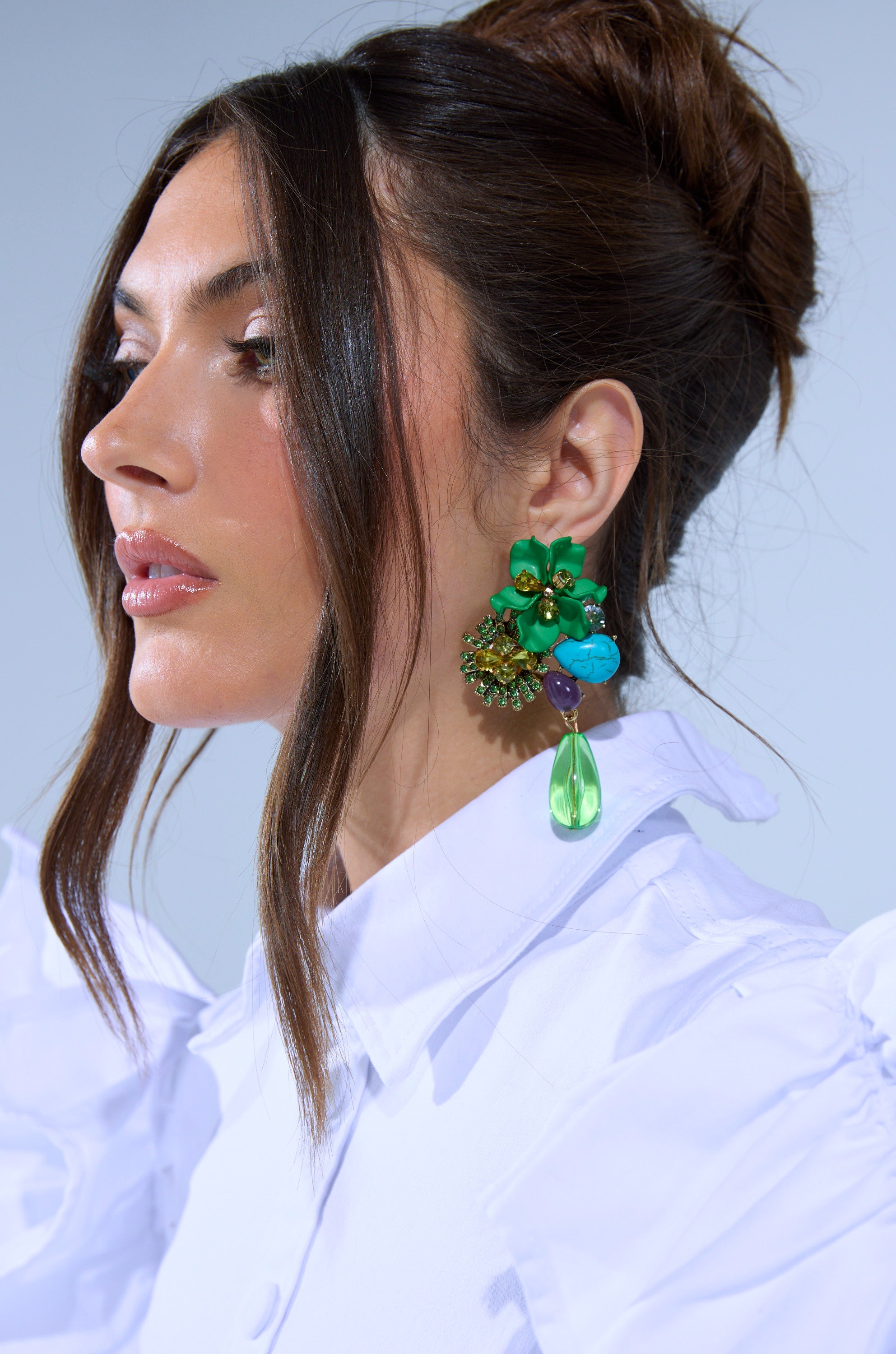 A woman in a white ruffled blouse and elegant updo models the GREEN GLORY EARRING, a floral design with green, blue, and gold hues, crystal accents, and a bold green teardrop pendant. 1