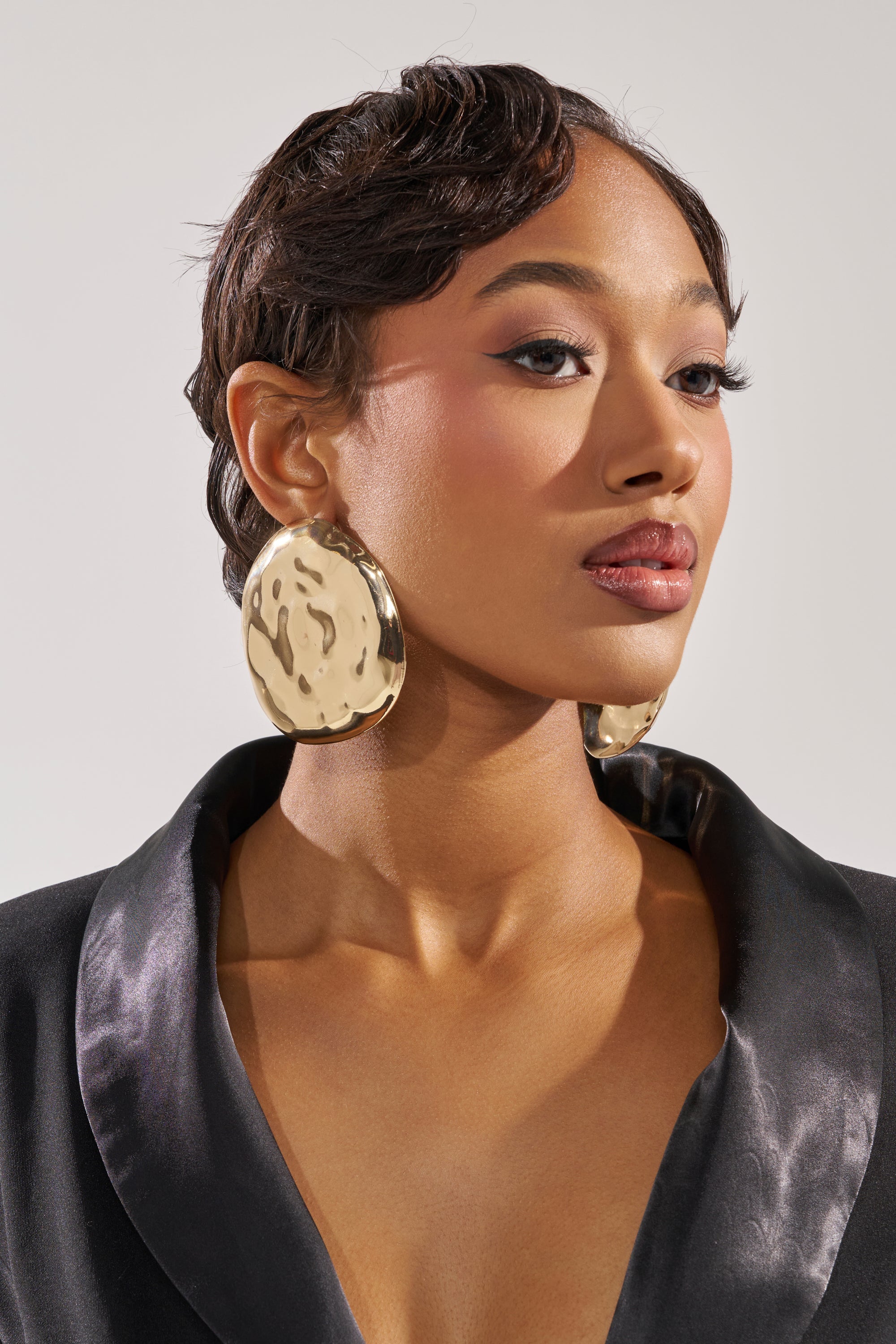A person with short, wavy hair wears a BENT OUTTA SHAPE EARRING—oversized and textured in hammered gold—paired with a silky black jacket. Their makeup features winged eyeliner and neutral lipstick as they gaze confidently to the side. 0