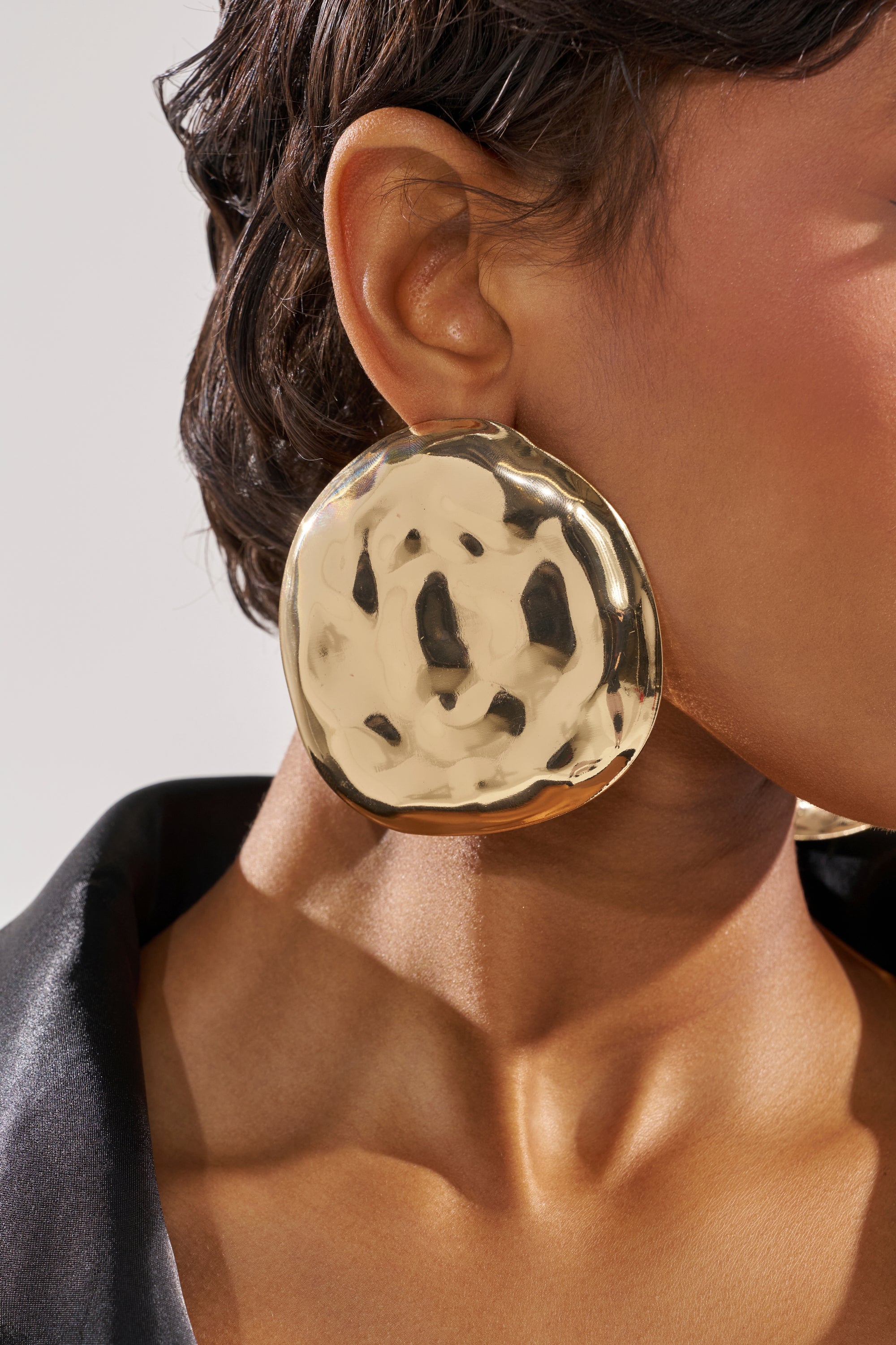 Close-up of a person wearing the BENT OUTTA SHAPE EARRING. The oversized, circular hammered gold earring has a shiny, textured surface. The person has short dark hair and is wearing part of a black garment. 1