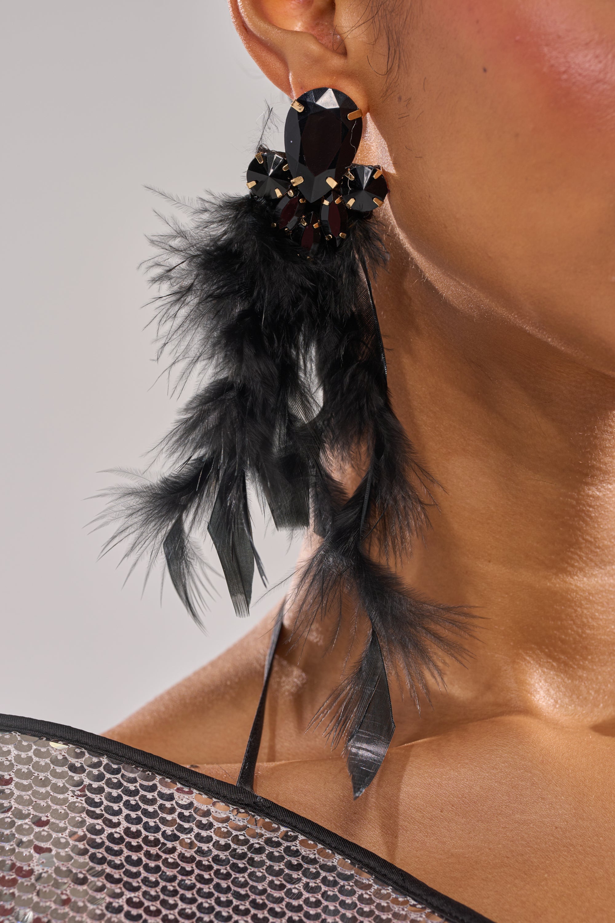 Close-up of a person wearing the SHOW ME EARRING—bold black feather earrings with large gemstones, reminiscent of glamorous showgirl style. Part of their cheek and shoulder is seen against a soft, neutral background. 1