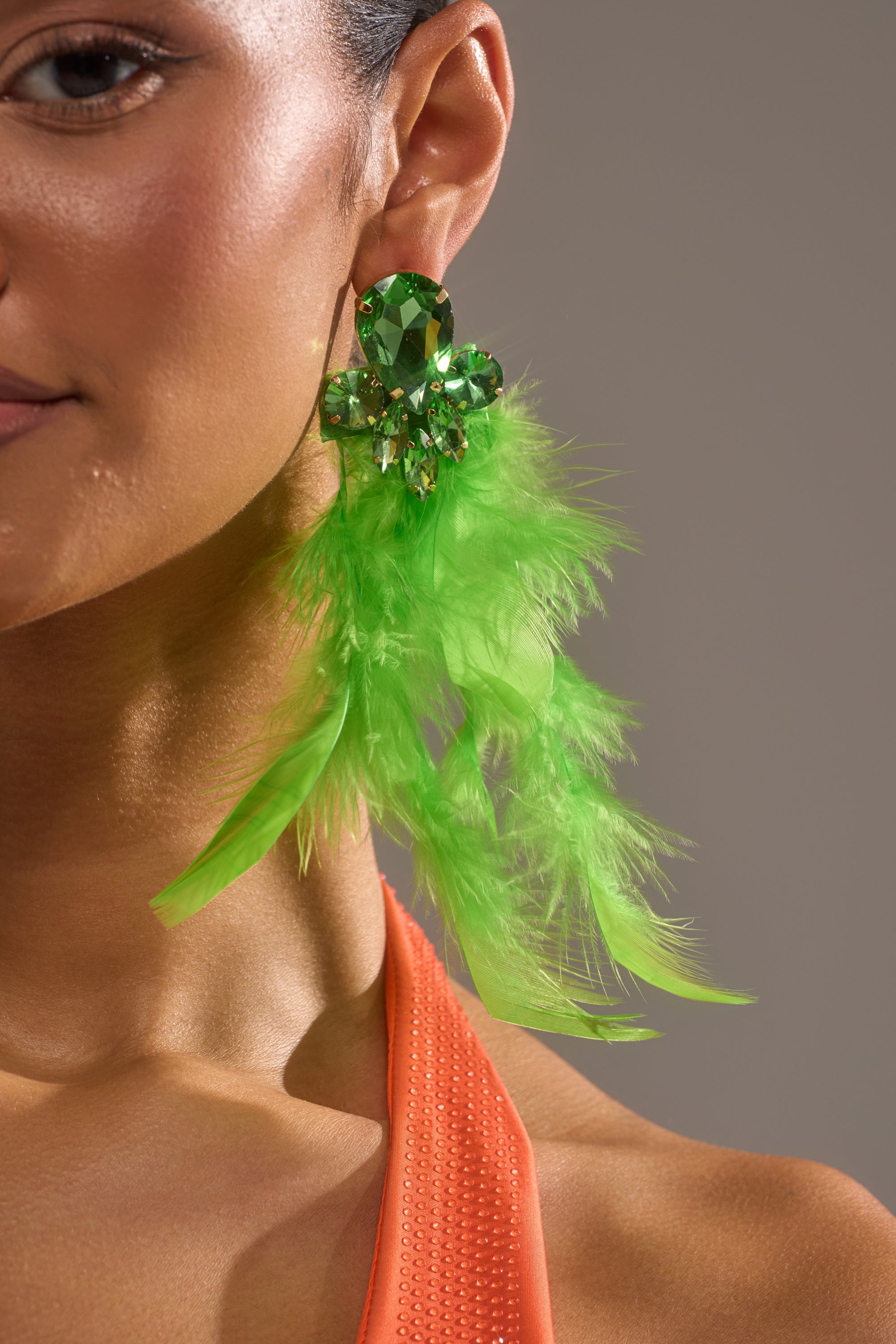 Close-up of a person wearing the SHOW IT OFF EARRING, featuring bold emerald-green feathers and sparkling gemstones. They have smooth skin and are dressed in an orange textured top. 0