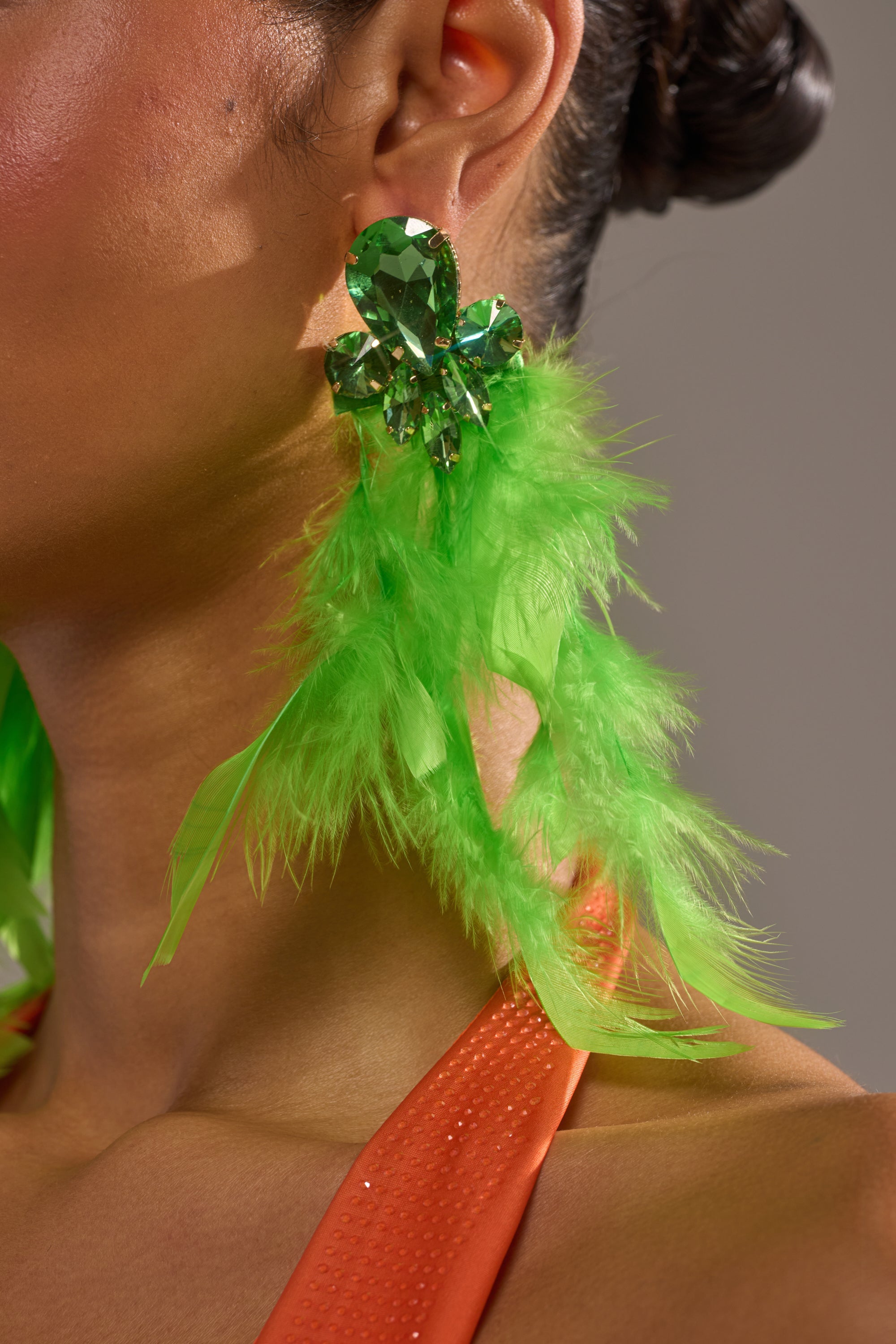 Close-up of a person wearing the SHOW IT OFF EARRING, featuring emerald-green crystal gems and neon green feather accents, boldly paired with an orange shimmery-textured top. 1