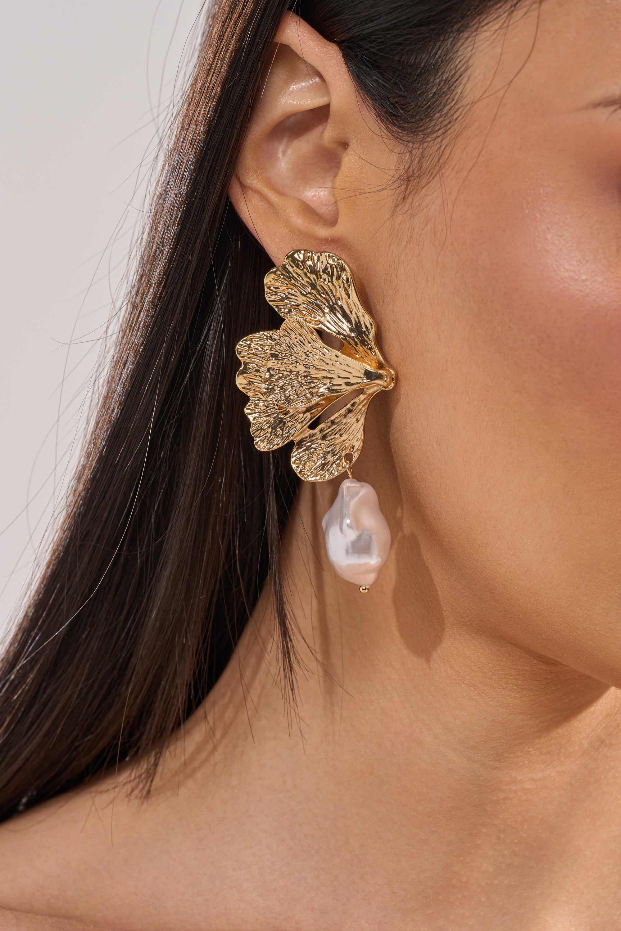 Close-up of a woman with straight dark hair and tanned skin wearing ON THE BEACH EARRING—gold-tone, leaf-shaped statement earrings featuring a dangling faux pearl. 2