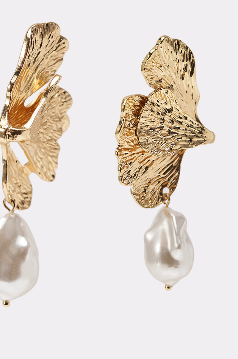 Close-up of ON THE BEACH EARRING: two gold-tone earrings shaped like textured leaves, each with a large, irregular white faux pearl drop. Set against a plain, light-colored background. 4