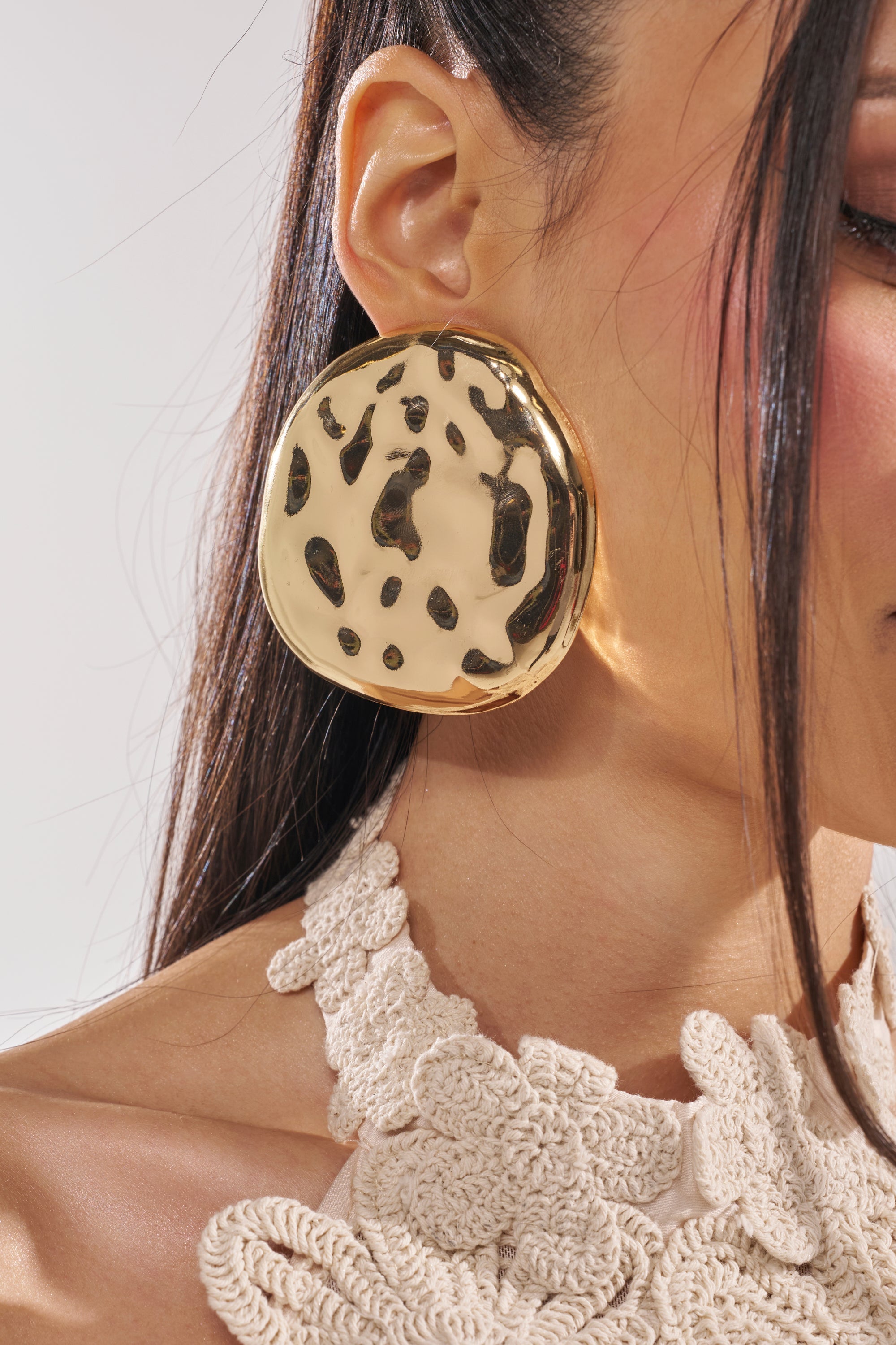 A close-up of a woman with straight dark hair wearing the CRUSHING IT EARRING—an oversized disc statement earring featuring black spots and a hammered finish. She pairs it with a cream crocheted textured top. 1