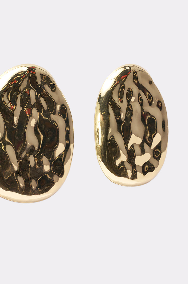 The CRUSHING IT EARRING features two shiny, irregularly shaped gold statement earrings with a hammered, textured surface displayed on a plain light background. 3