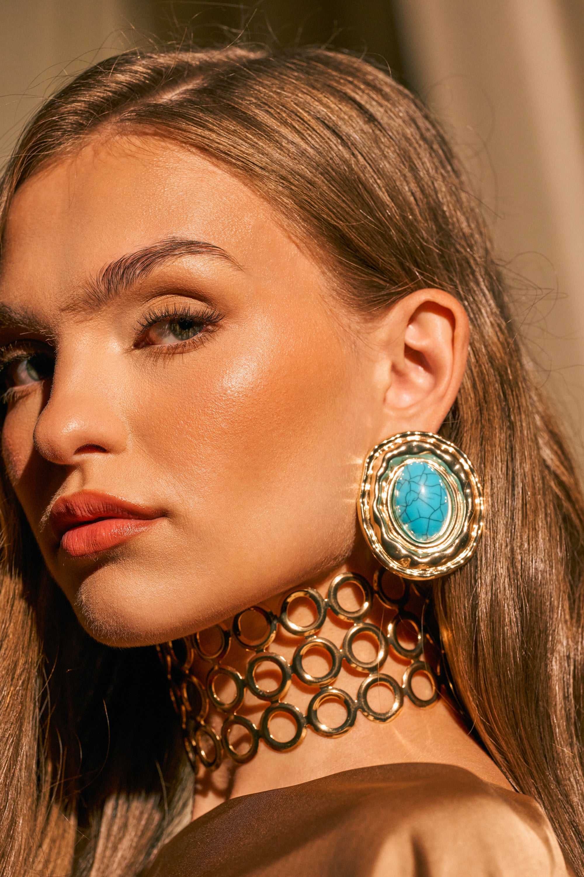 A woman with smooth brown hair wears the LONDON EARRING, an oversized oval stud featuring a turquoise center stone, paired with a gold choker adorned with circular links. She showcases a natural makeup look and is dressed in neutral tones.