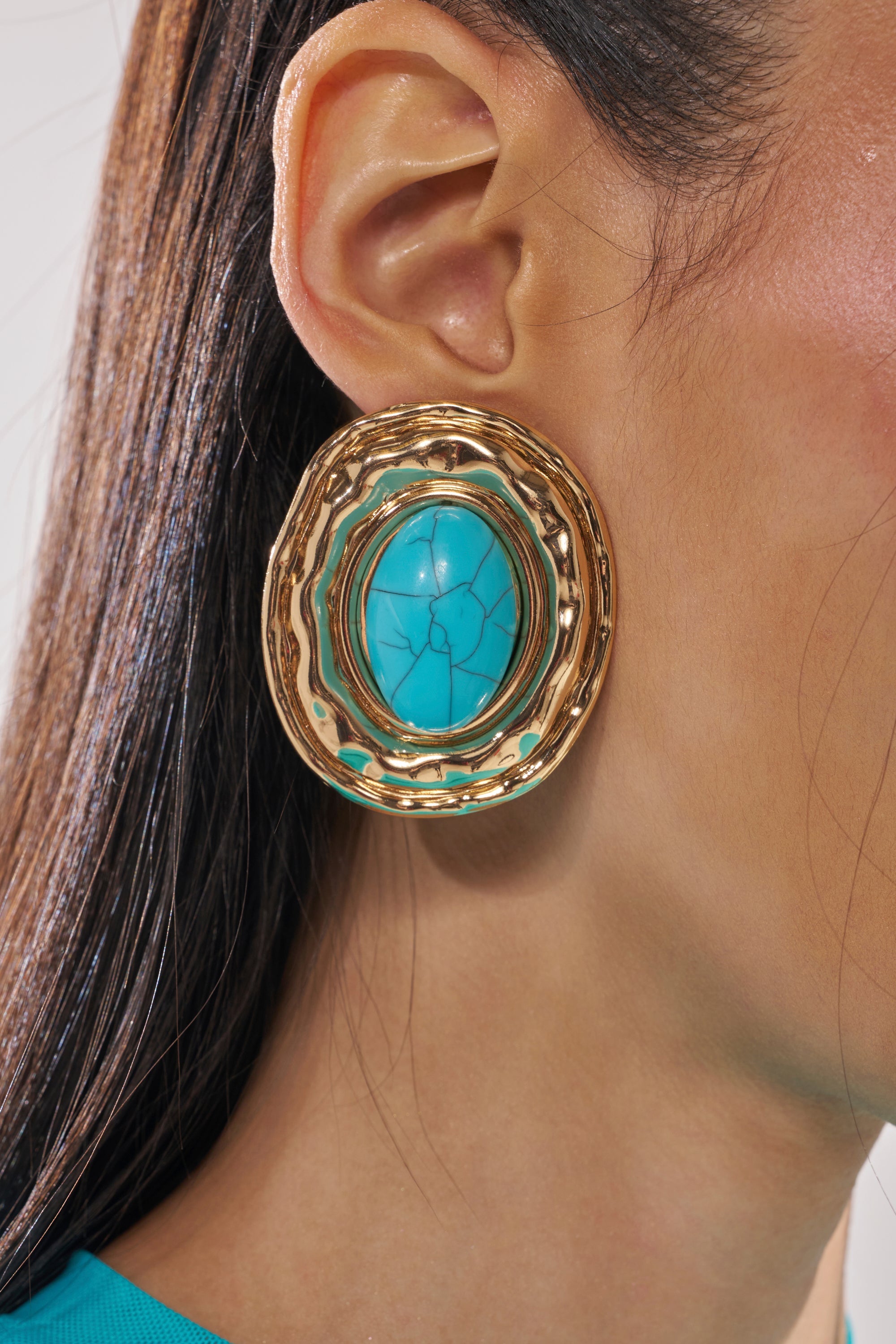 Close-up of a person wearing the LONDON EARRING—oversized ovals with a textured gold border and turquoise stone center. The person has straight brown hair and is dressed in a teal top. 4