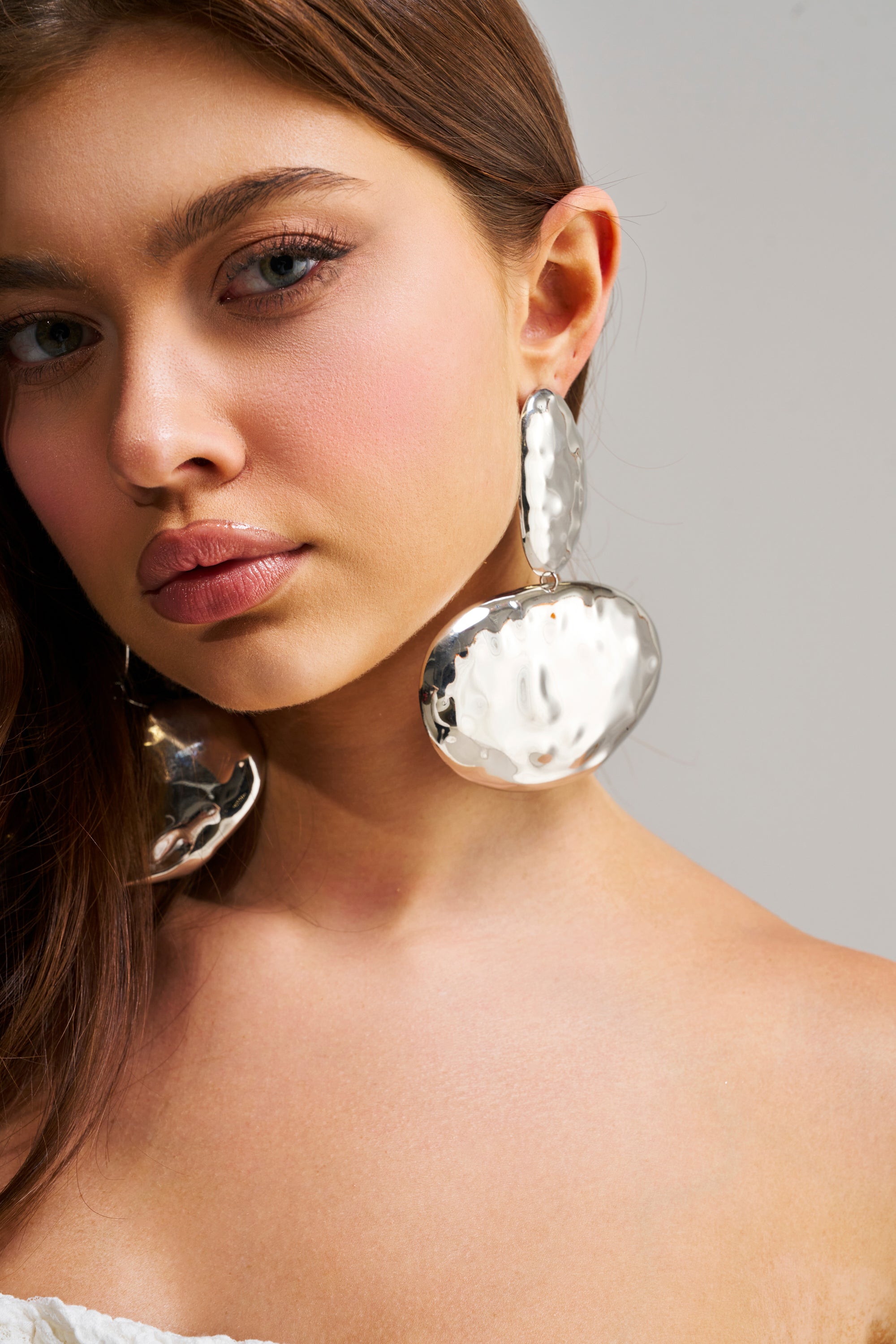 A woman with long brown hair models the ORGANIC VIBES EARRING—oversized circular silver statement earrings—with a white off-shoulder top, posing against a plain light background.