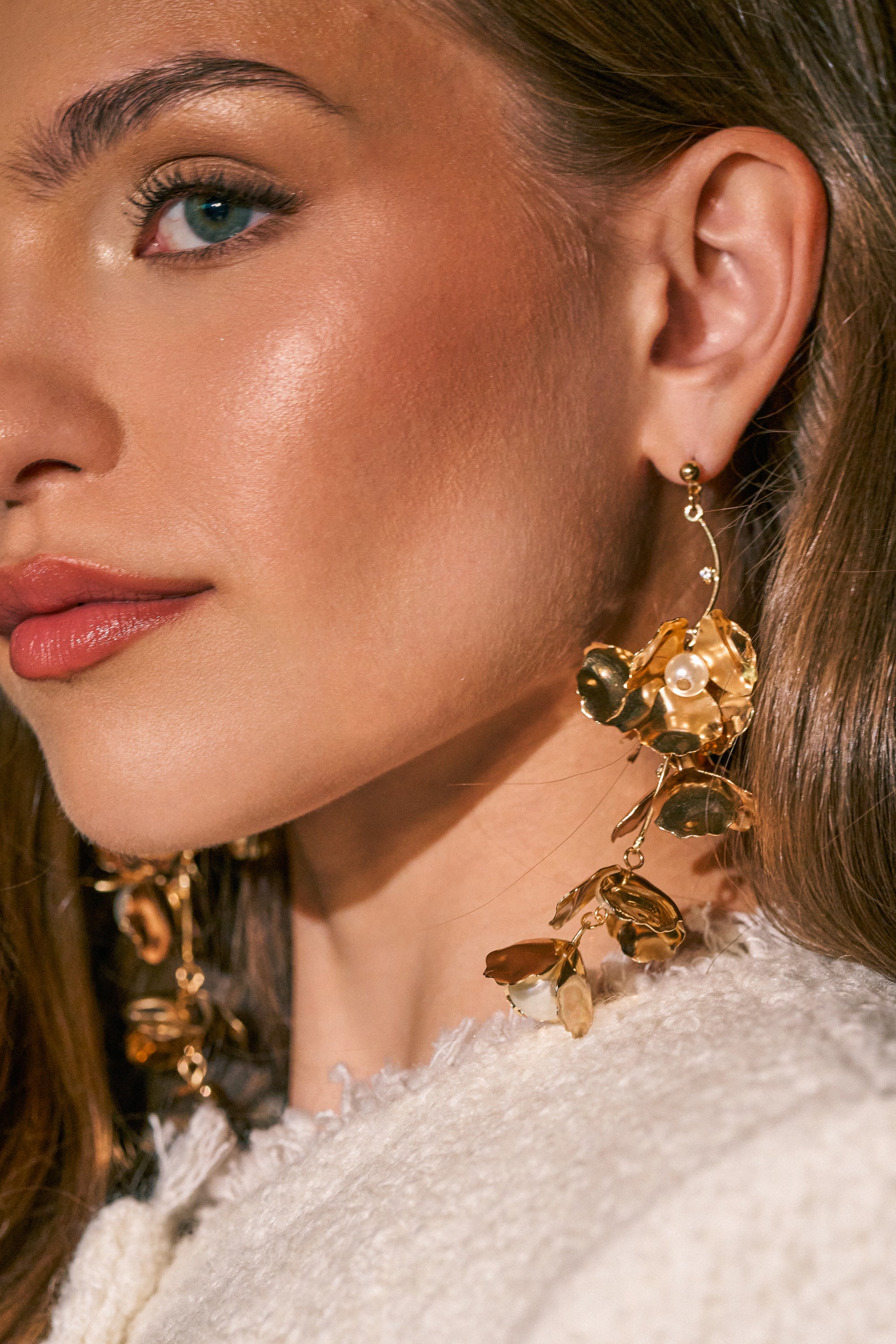 A close-up of a woman with long brown hair wearing the NOT THE NORM EARRING—elegant gold drop earrings featuring a floral design—paired with a cream textured top. Her natural makeup enhances her blue eyes and pink lips, creating an effortlessly fresh look that perfectly highlights these spring-ready earrings.