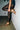 Wearing AZALEA WANG YEIRA GOLD SANDAL with chunky chain links and comfort cushion insoles, a person in black lace leggings leans against a white brick wall, one foot raised and both hands holding their ankle, lower body only visible.