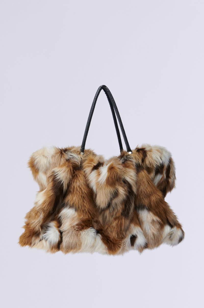 The UPSETTER FAUX FUR TOTE is an oversized handbag made from shagadelic faux fur in a luxurious mix of brown, white, and black colors, accented with sleek black leather handles, showcased against a simple light gray background.