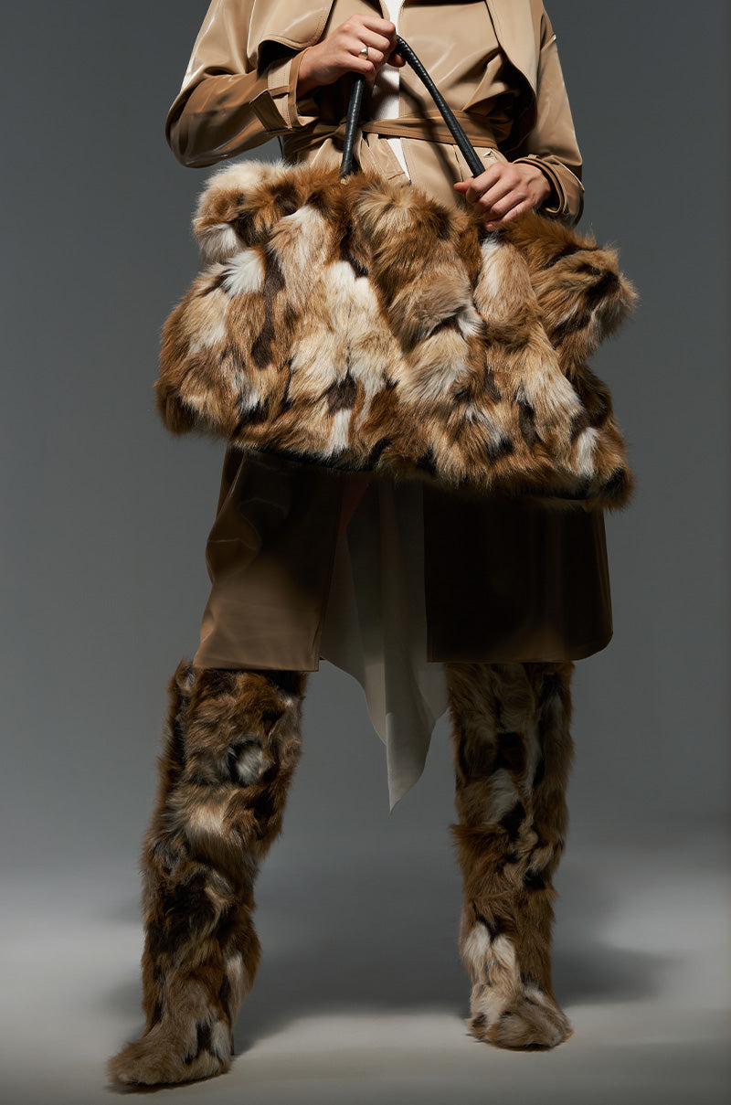 Dressed in a brown ensemble, an individual carries the UPSETTER FAUX FUR TOTE, which features a large, shagadelic design in brown and white faux fur. Complementary tall, furry boots complete the look against a neutral gray background. 0