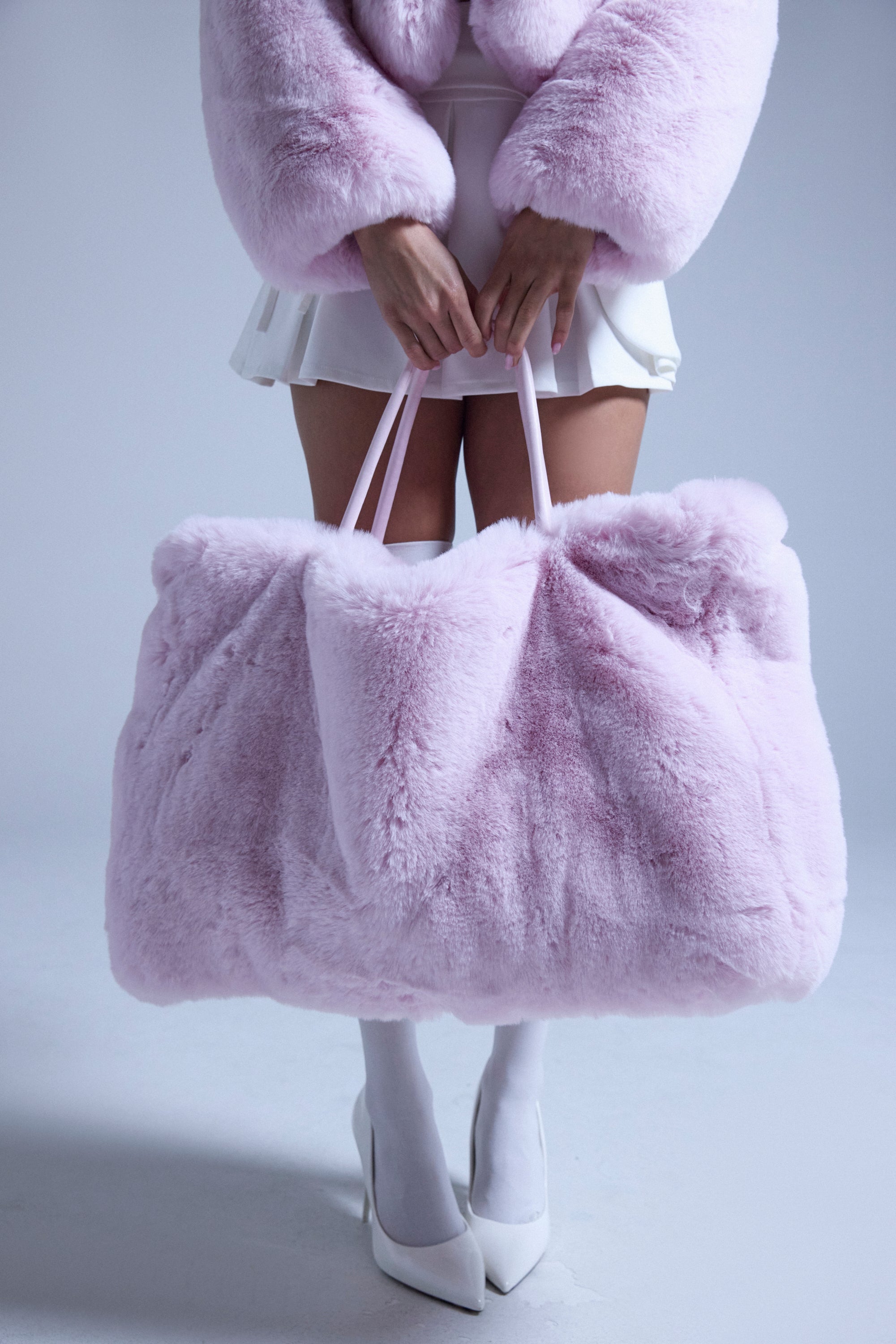 A person holds the BB GURLLLL FAUX FUR TOTE, an oversized faux fur bag with a satin-lined interior, while wearing a pink faux fur jacket, white skirt, tights, and heels against a light gray background. 1