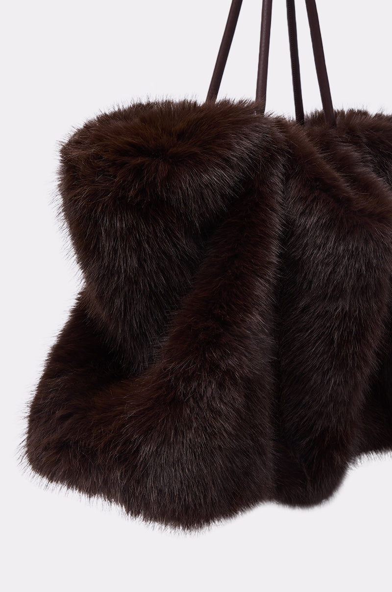 A close-up of the SO RICH FAUX FUR TOTE shows its dark brown, fluffy faux fur and long black leather straps against a light background, highlighting the bag’s soft texture and rich color.