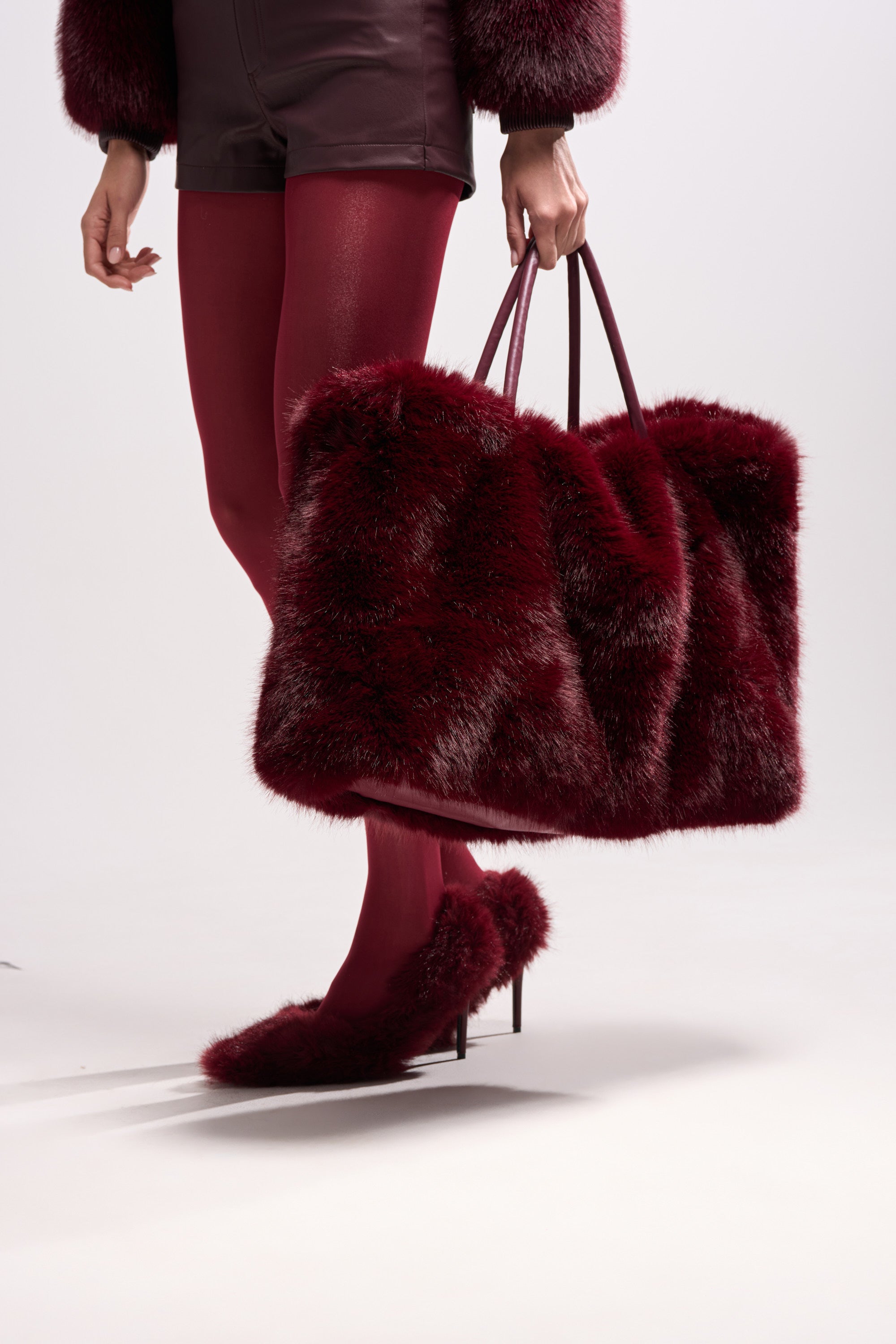 A person carries the ALAIA FAUX FUR TOTE, paired with wine-red faux fur shoes, shorts, and tights. Part of their upper body is out of frame, set against a plain light background.