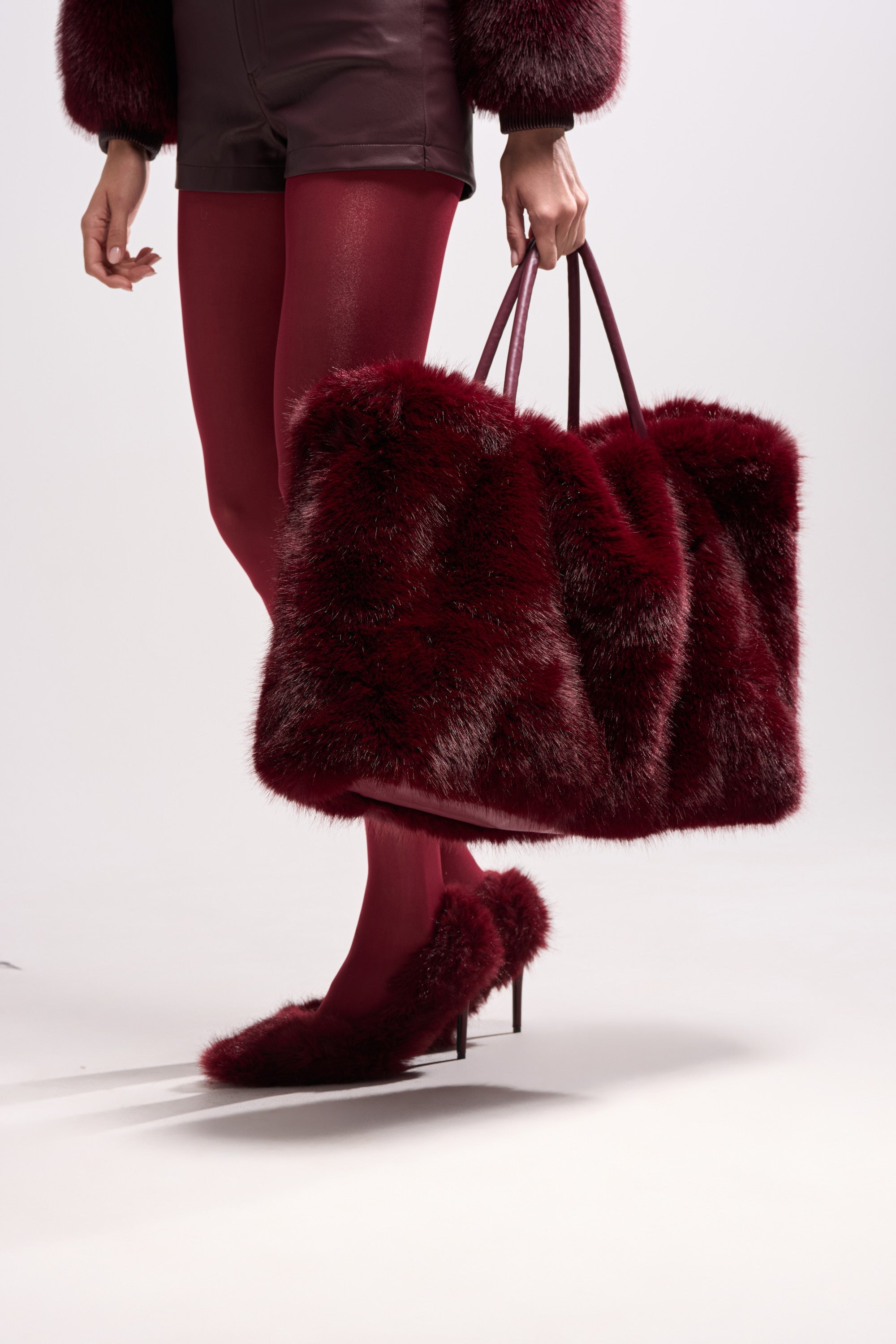 A person carries the ALAIA FAUX FUR TOTE, paired with wine-red faux fur shoes, shorts, and tights. Part of their upper body is out of frame, set against a plain light background.