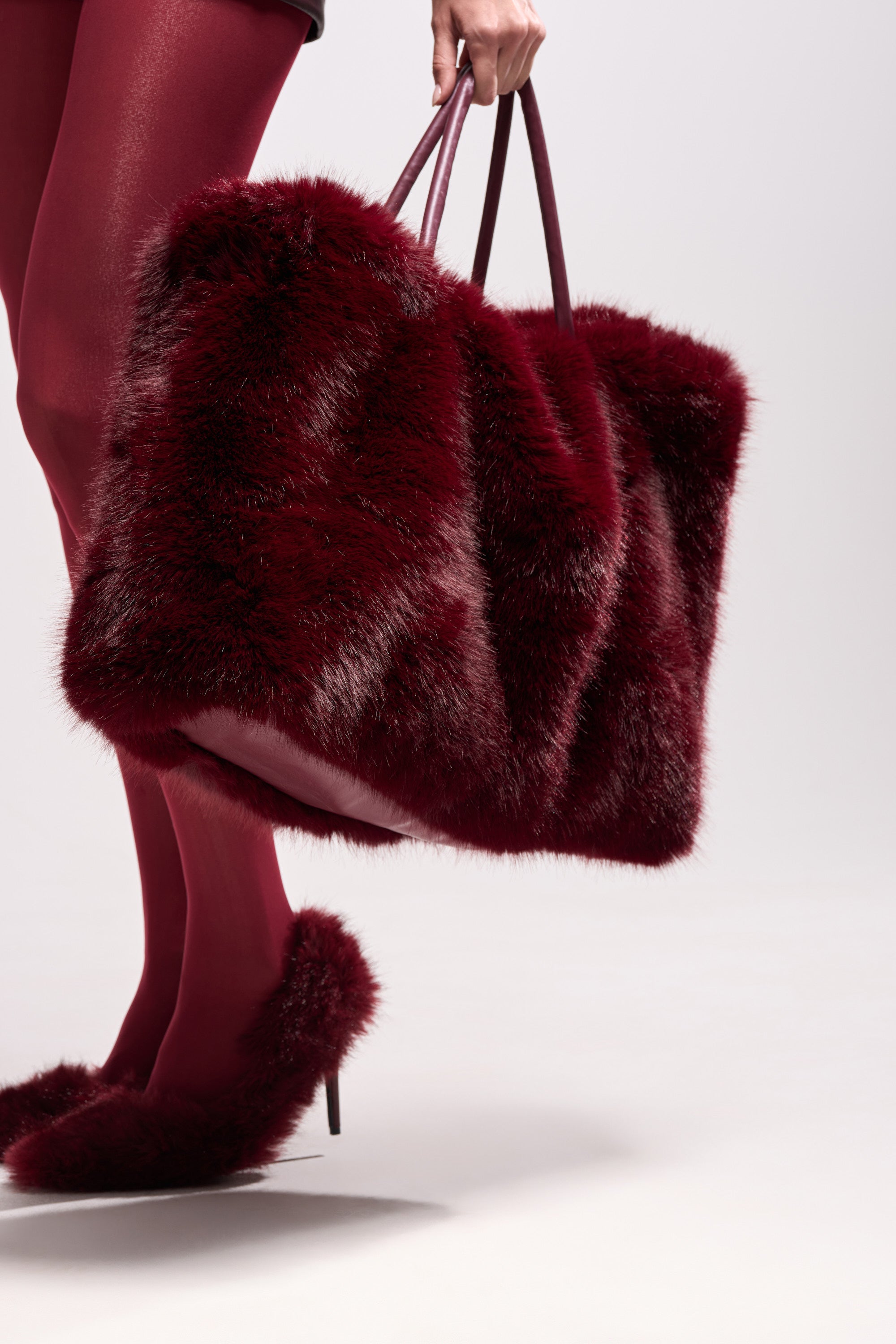 Wearing deep red tights and furry heels, a person carries the oversized ALAIA FAUX FUR TOTE in wine-red. A plain, light background highlights the tote's rich texture and statement style.