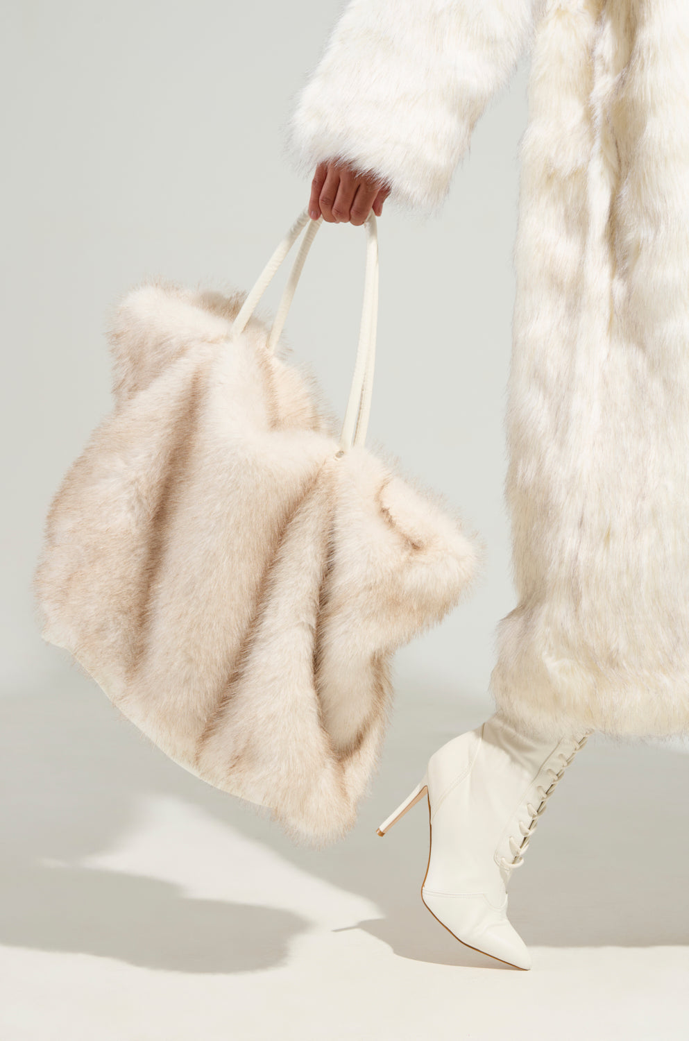 A person in a white faux fur coat and white lace-up high-heeled boots carries an oversized, fluffy TOASTIE FAUX FUR TOTE in beige. The background is a plain, light-colored surface.