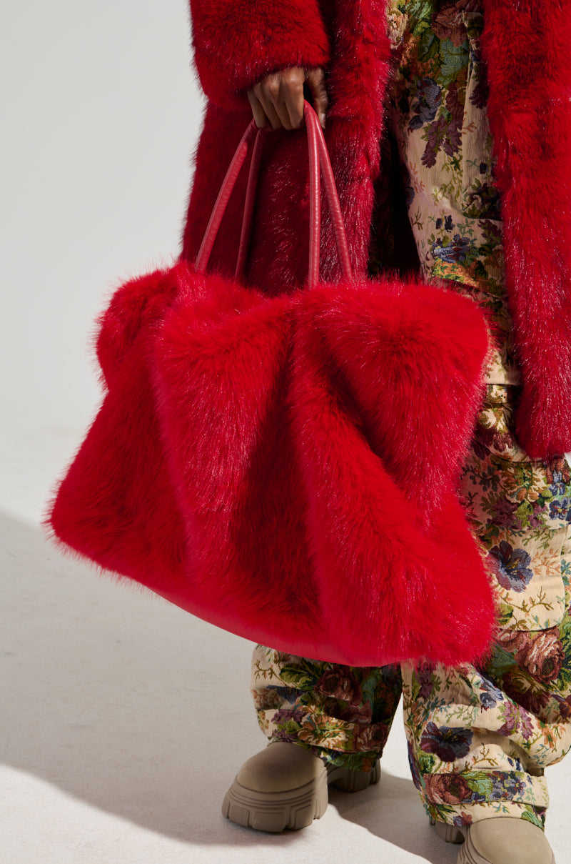 A person wearing a shagadelic faux fur red coat holds the LOOK AT ME NOW FAUX FUR TOTE, an oversized handbag. They are dressed in floral-patterned pants and beige boots, set against a plain, light-colored backdrop. 0