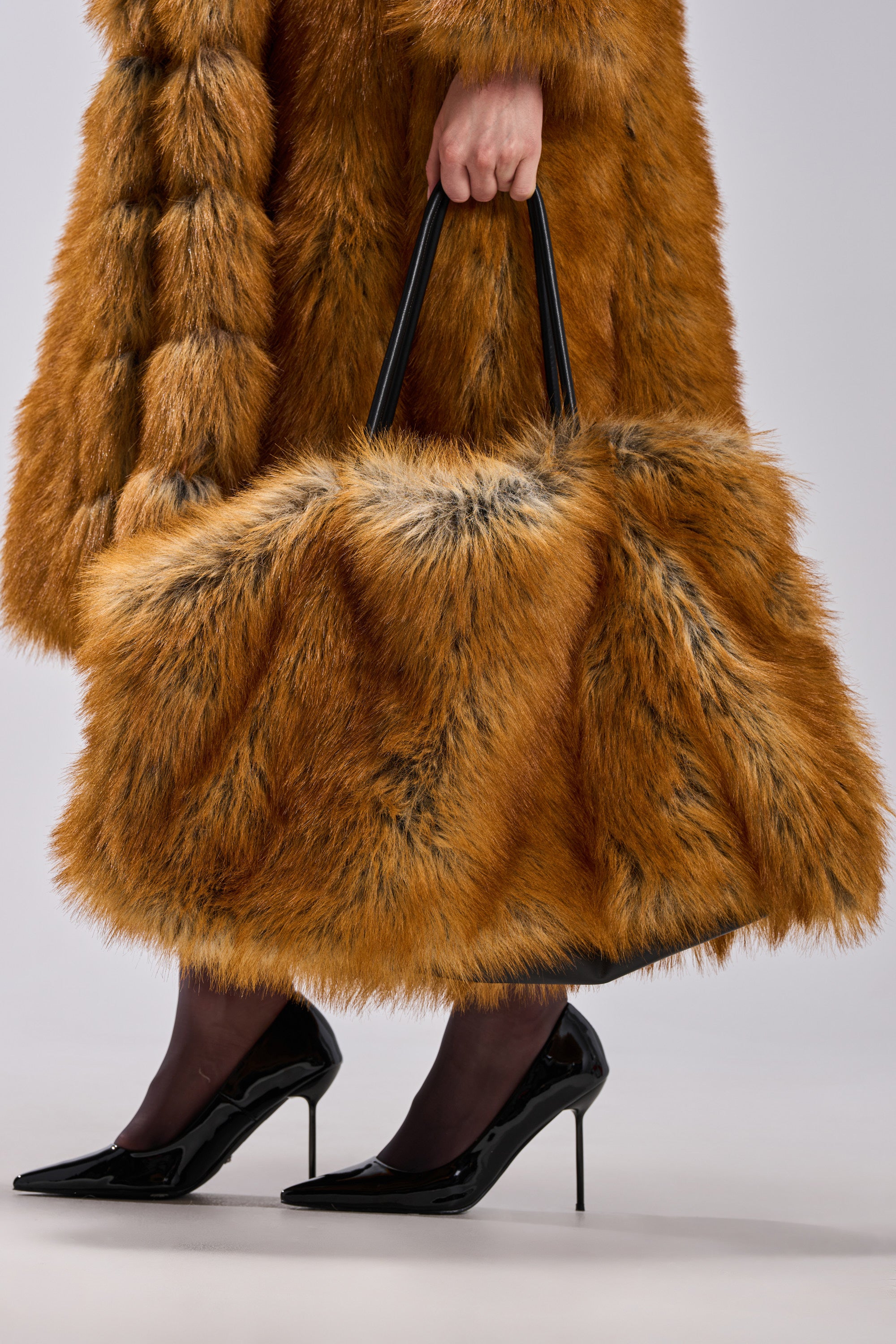 A person in a brown faux fur coat and black heels carries the HERE FOR A GOOD TIME FAUX FUR TOTE, a large brown bag with black handles, against a plain neutral background.