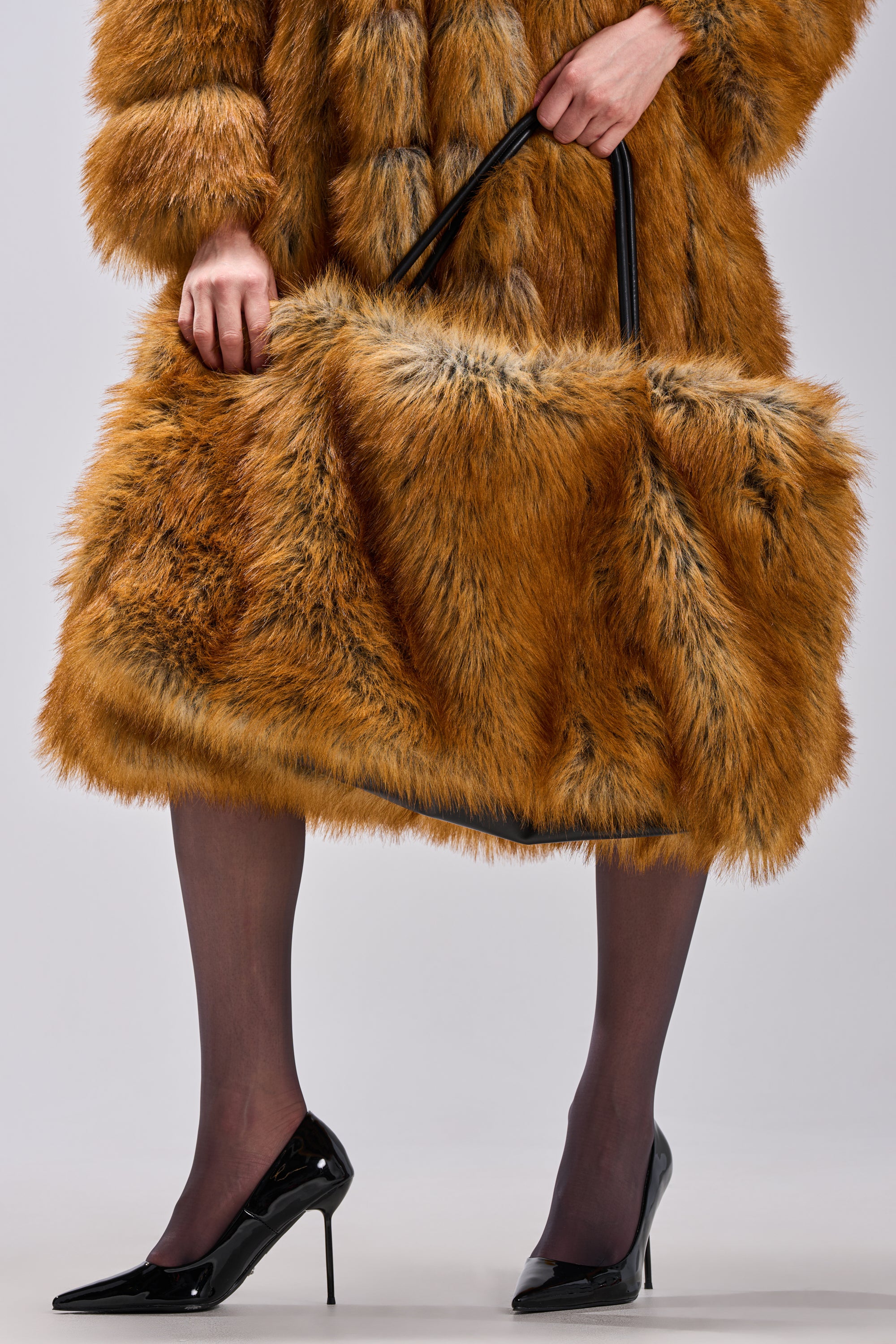 A person in a brown fur coat and sheer black tights holds the HERE FOR A GOOD TIME FAUX FUR TOTE. They also wear black heels, with only the lower half of their body visible.