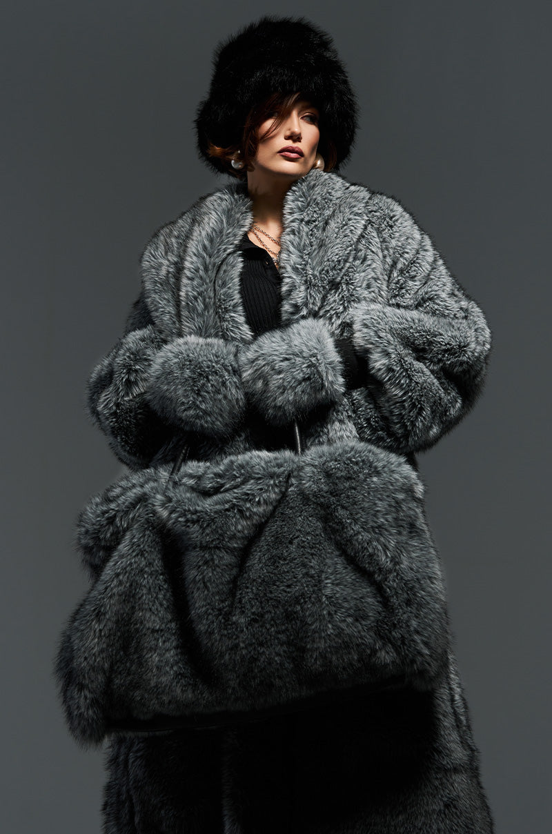 A person clad in a luxurious, oversized shagadelic faux fur gray coat and a black fur hat stands against a gray backdrop. With their hands tucked into the coat's pockets, they exude confidence, enhanced by the stylish TSUNAMI FAUX FUR TOTE that complements their chic ensemble.