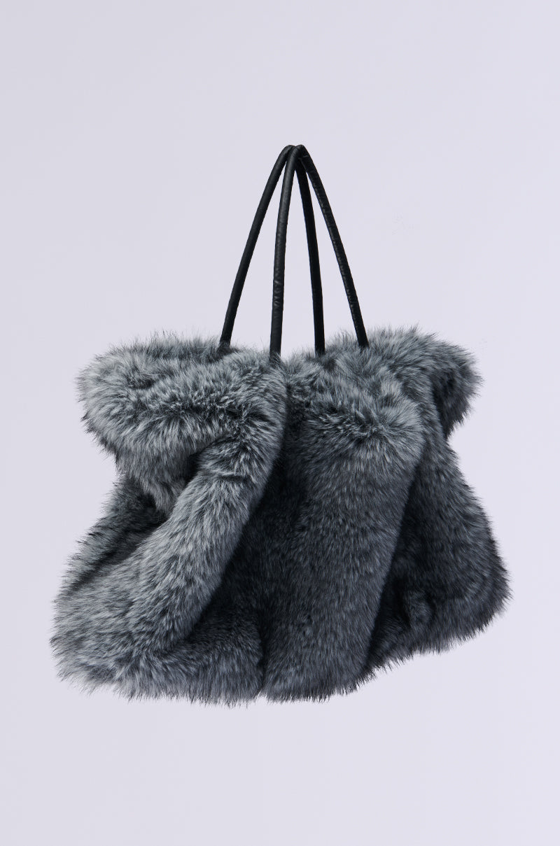 The TSUNAMI FAUX FUR TOTE is highlighted against a minimalistic light background, featuring an oversized design in grey shagadelic faux fur and accented by two sleek black handles. Its plush and voluminous form radiates a sense of comfort and luxury.