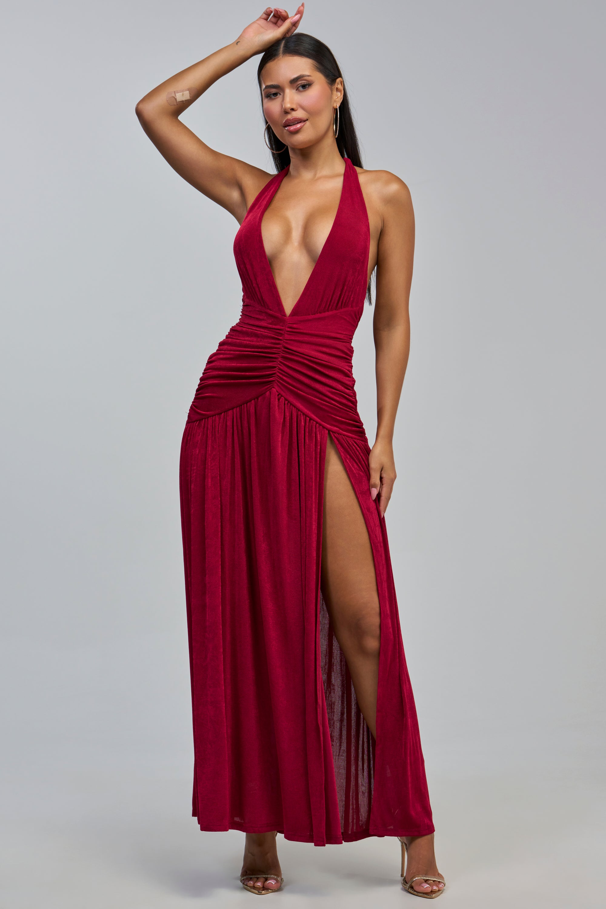 Wearing the POISON IVY MAXI DRESS IN BURGUNDY, a woman stands confidently in strappy heels against a plain light gray background. The striking maxi dress features a halter neckline, plunging front, ruched waist, and a daring high slit that reveals one leg.