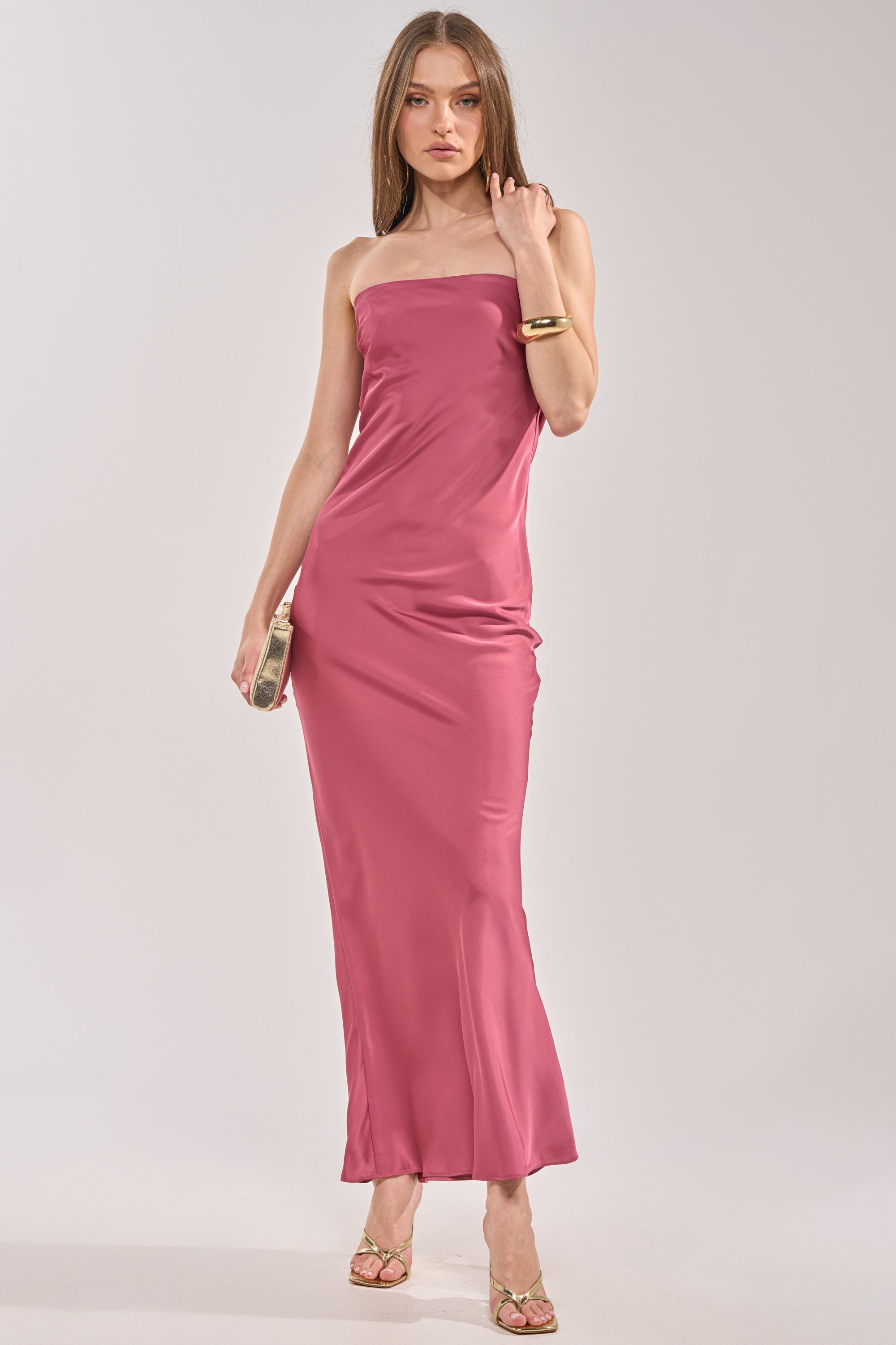 A woman poses gracefully in the SATIN SINNER MAXI DRESS—pink satin and strapless—paired with gold high-heeled sandals and a metallic clutch, creating an elegant wedding guest look against a plain light background. 1