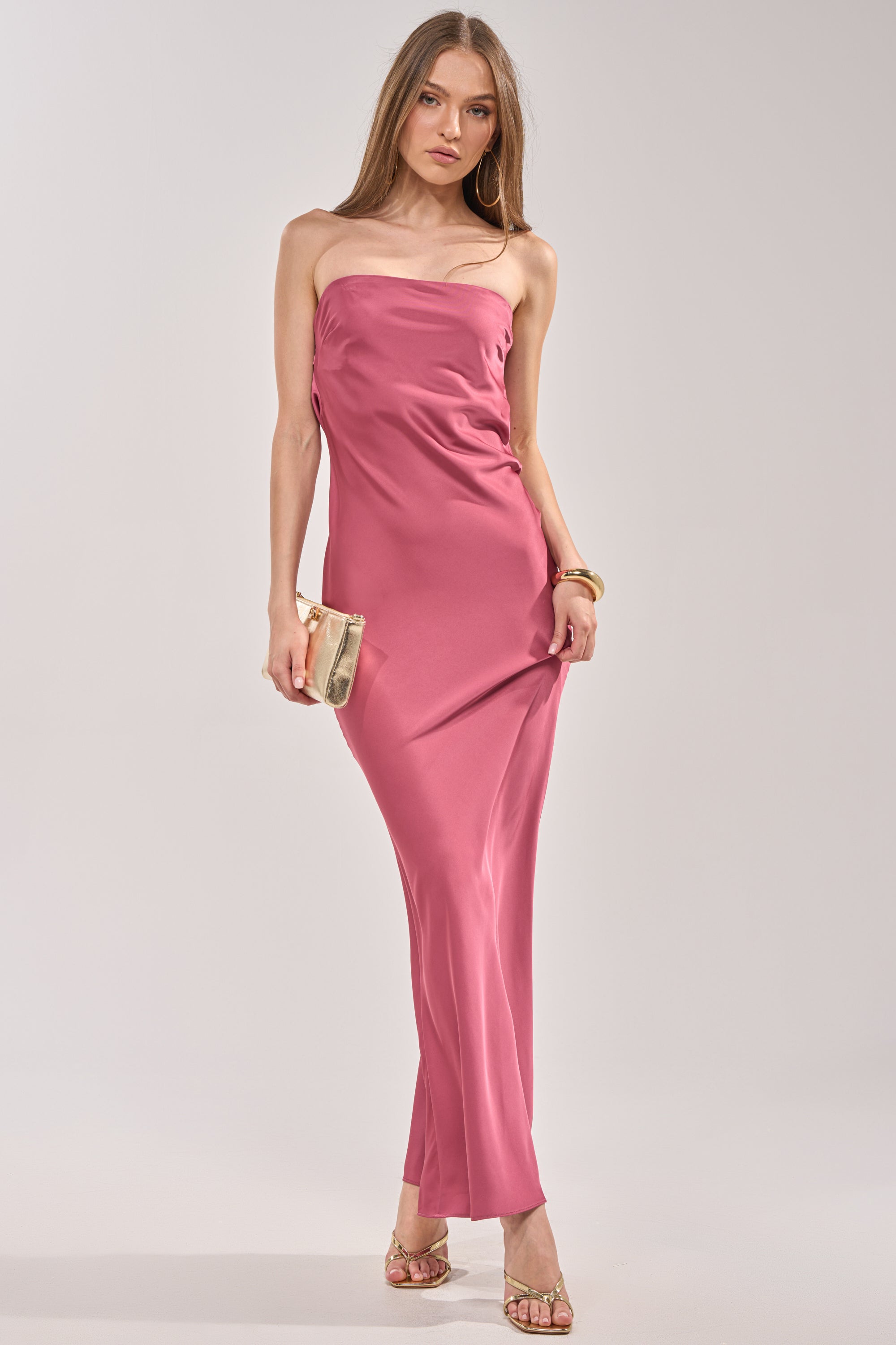 She models the SATIN SINNER MAXI DRESS, a strapless pink satin maxi styled with a gold clutch, hoop earrings, a bracelet, and heeled sandals—ideal for wedding guests—posing confidently against a plain backdrop. 2