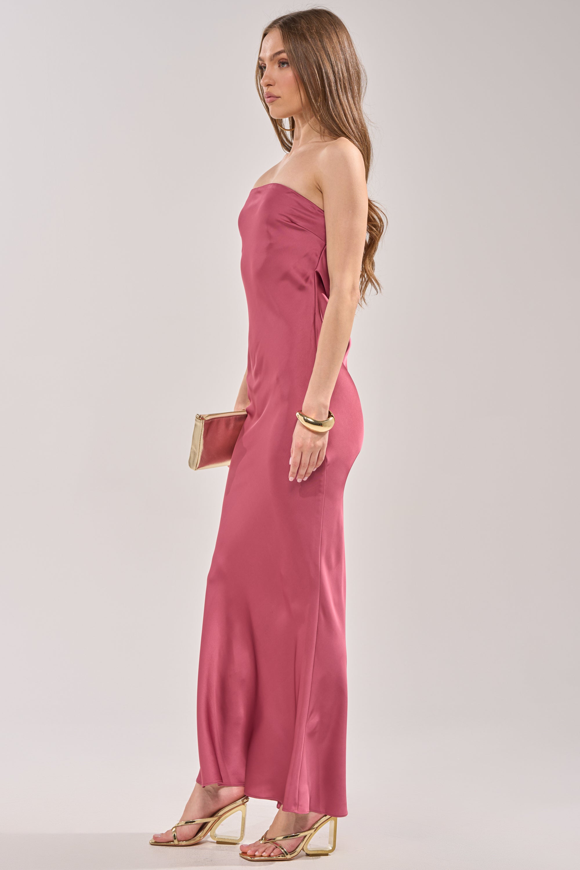 A woman in profile wears the SATIN SINNER MAXI DRESS, a strapless pink satin maxi. She accessorizes with a beige clutch, gold bangles, and gold high-heeled sandals. Her long wavy brown hair completes this chic wedding guest look. 3