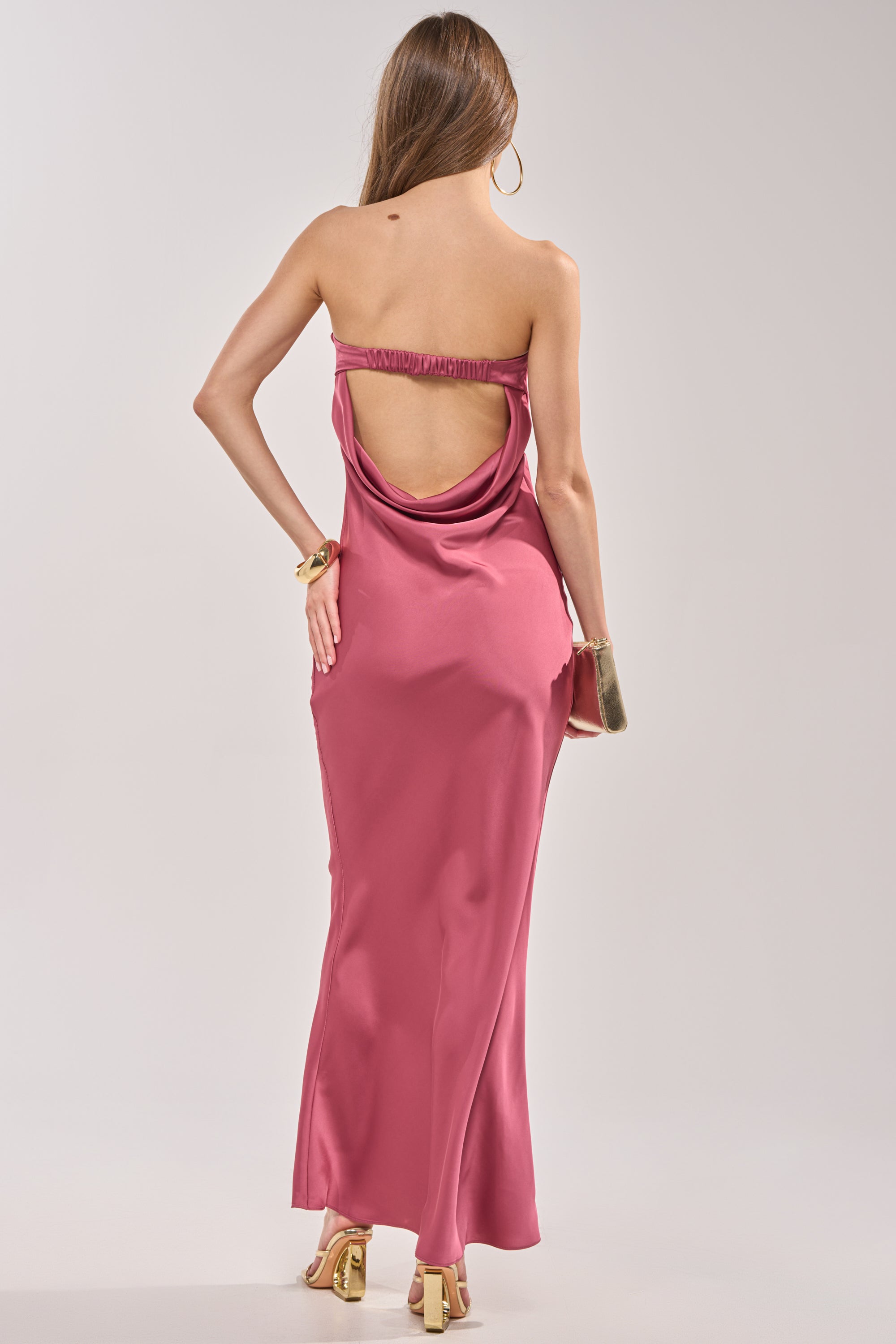 A woman with long brown hair wears the SATIN SINNER MAXI DRESS, a strapless pink satin maxi dress, styled with gold high heels, a gold clutch, hoop earrings, and a chunky bracelet for an ideal wedding guest look. 4