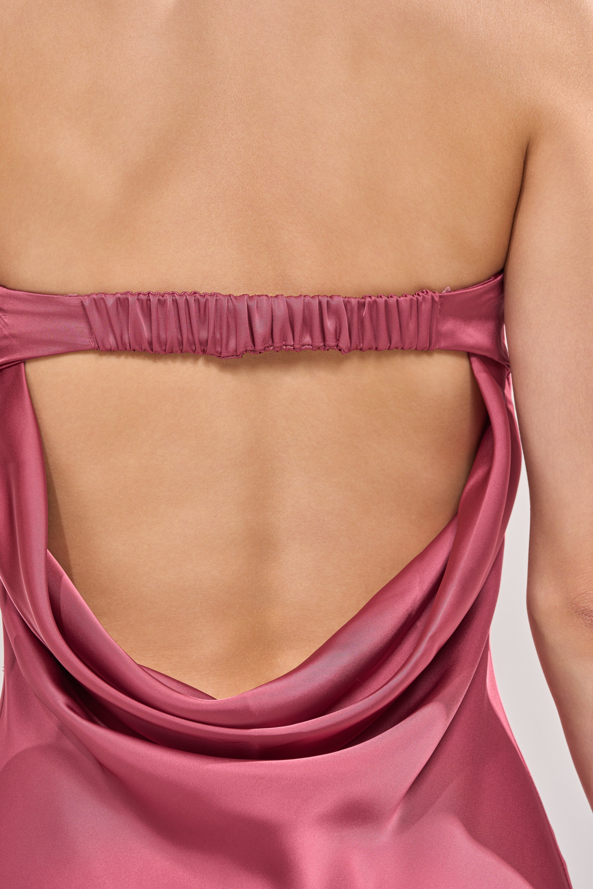 A close-up shows a woman's back in the SATIN SINNER MAXI DRESS, featuring pink satin fabric and a gathered upper back strap. The smooth, light-reflecting material makes this dress an elegant option for wedding guests. 5