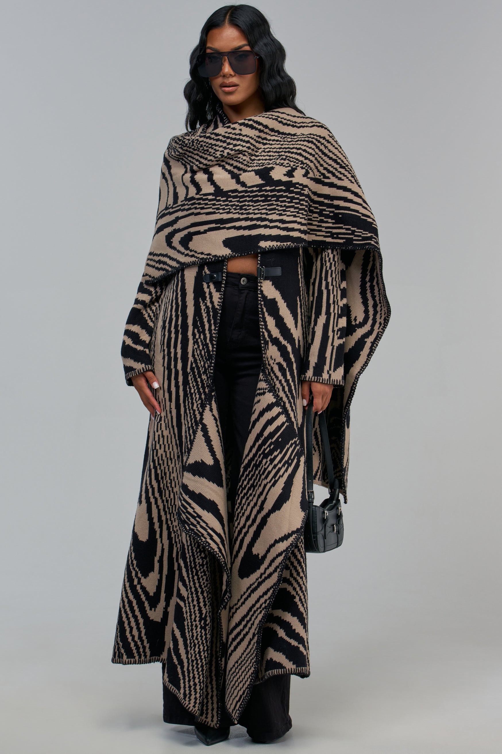 A person wearing a long, tan and black abstract WRAP ME UP KNIT CARDIGAN over black jeans and carrying a small black handbag.