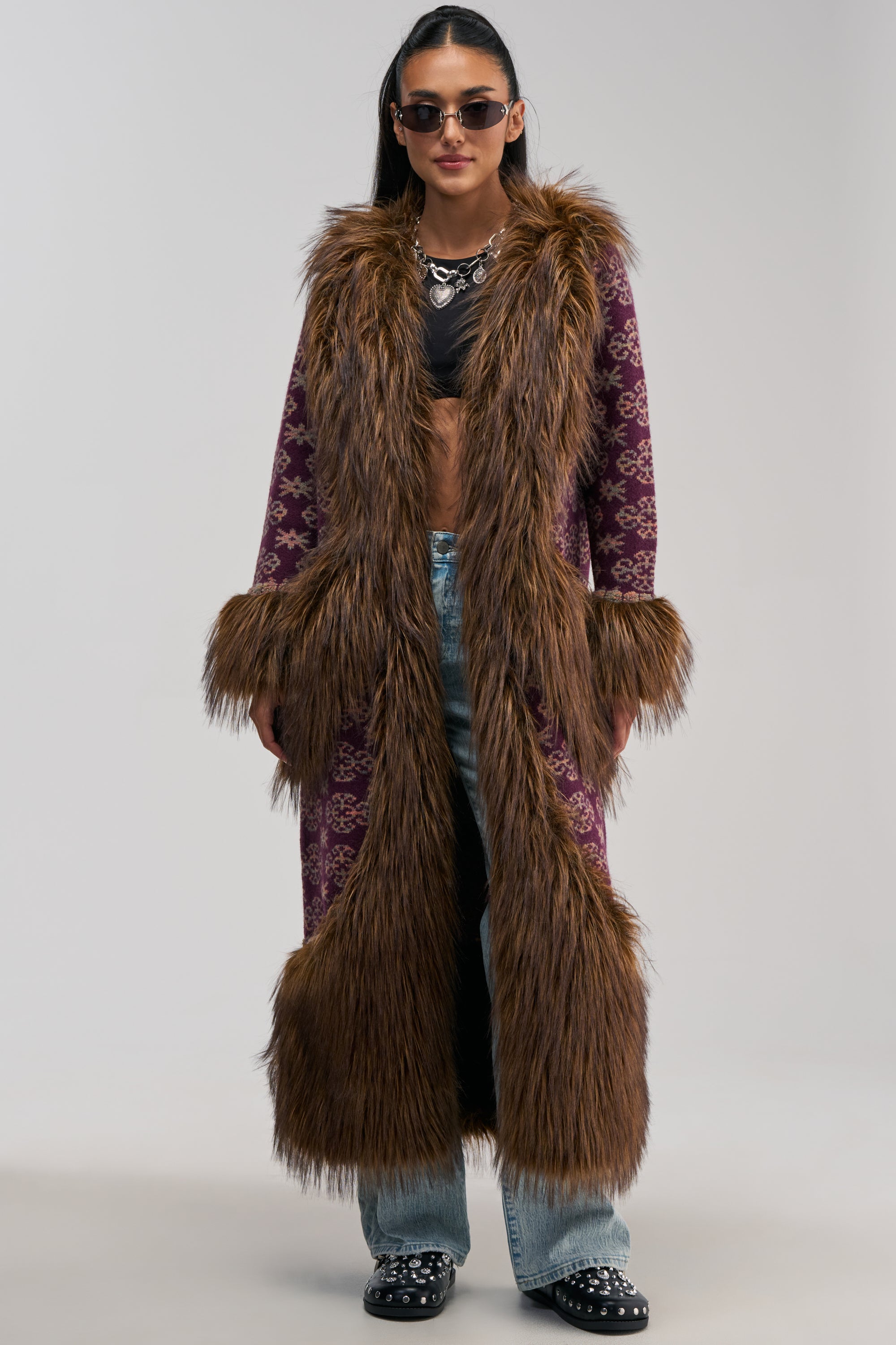 A person wearing the MAIN CHARACTER FUR TRIM CARDIGAN—a long, purple patterned coat with brown faux fur trim—styled with sunglasses, a black crop top, light blue jeans, black loafers, and a silver necklace against a plain background.
