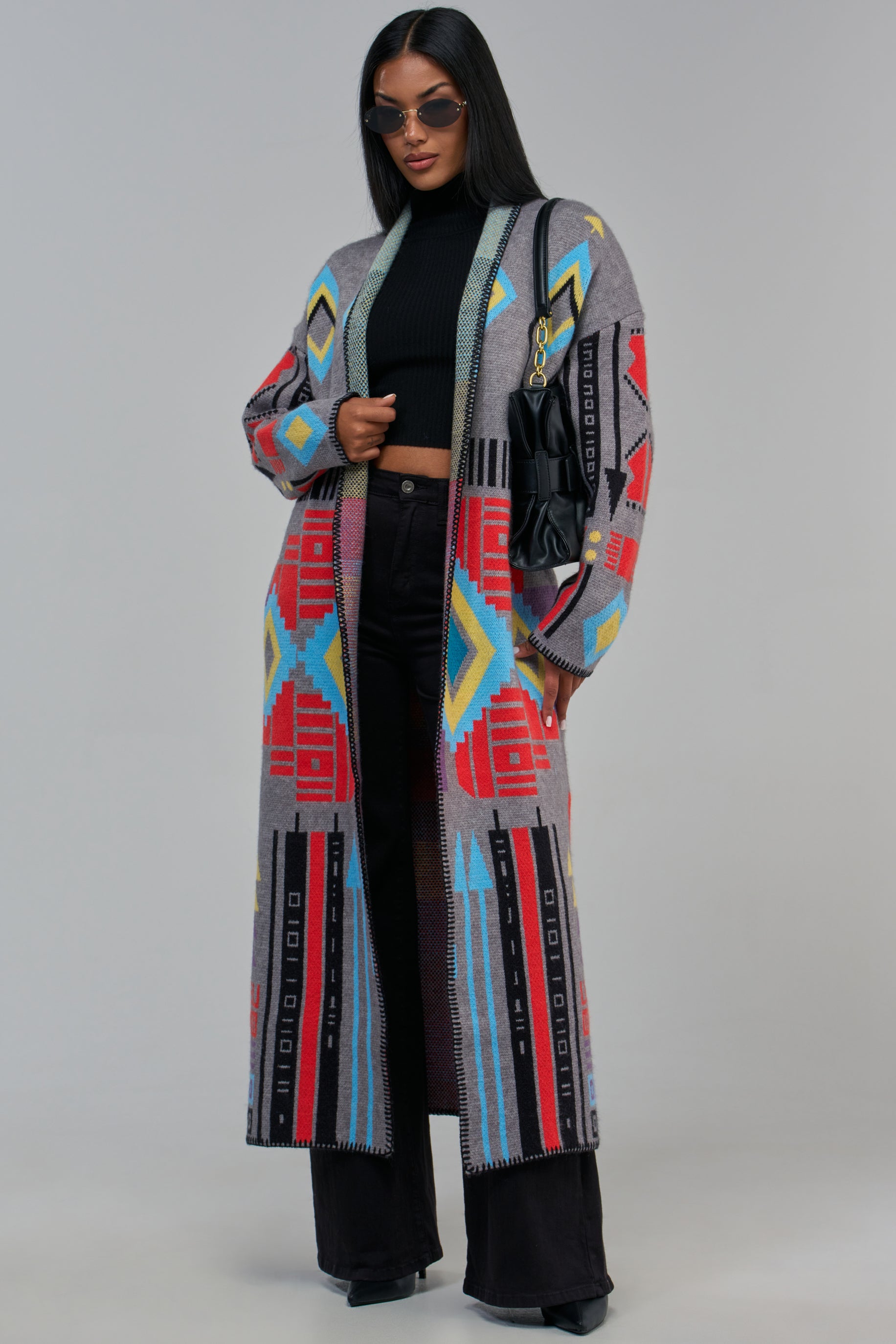 A model wears the LEAH KNIT CARDIGAN, a longline, gray open-front piece featuring a bold geometric pattern in red, blue, and yellow over a black top and black trousers.