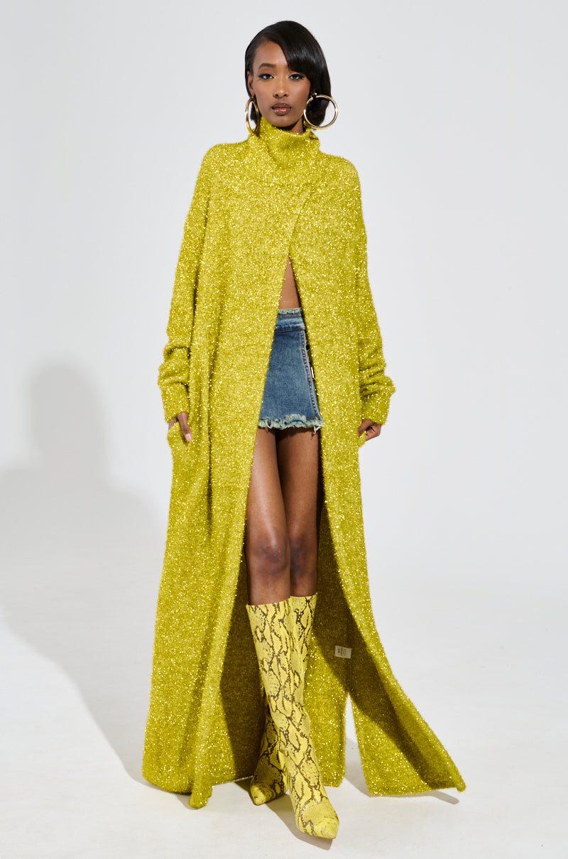 A woman poses effortlessly in the MOONLIGHT LONG TURTLENECK CARDIGAN IN GOLD, its shimmering open front revealing a chic denim mini skirt. She completes her look with knee-high yellow snakeskin boots and bold hoop earrings, radiating confidence against a minimalistic backdrop. Her style beautifully combines the cardigan's maxi length elegance with an urban edge.