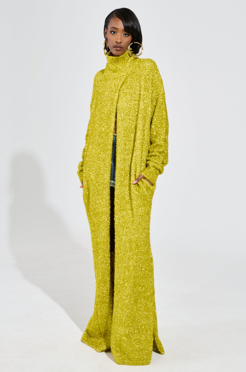 A woman models the MOONLIGHT LONG TURTLENECK CARDIGAN IN GOLD, showcasing its vibrant yellow hue against a plain white backdrop. The cardigan features a high collar and full sleeves, crafted from stretchy knit for ultimate comfort. With one hand slipped into a pocket, she completes her look with large hoop earrings.