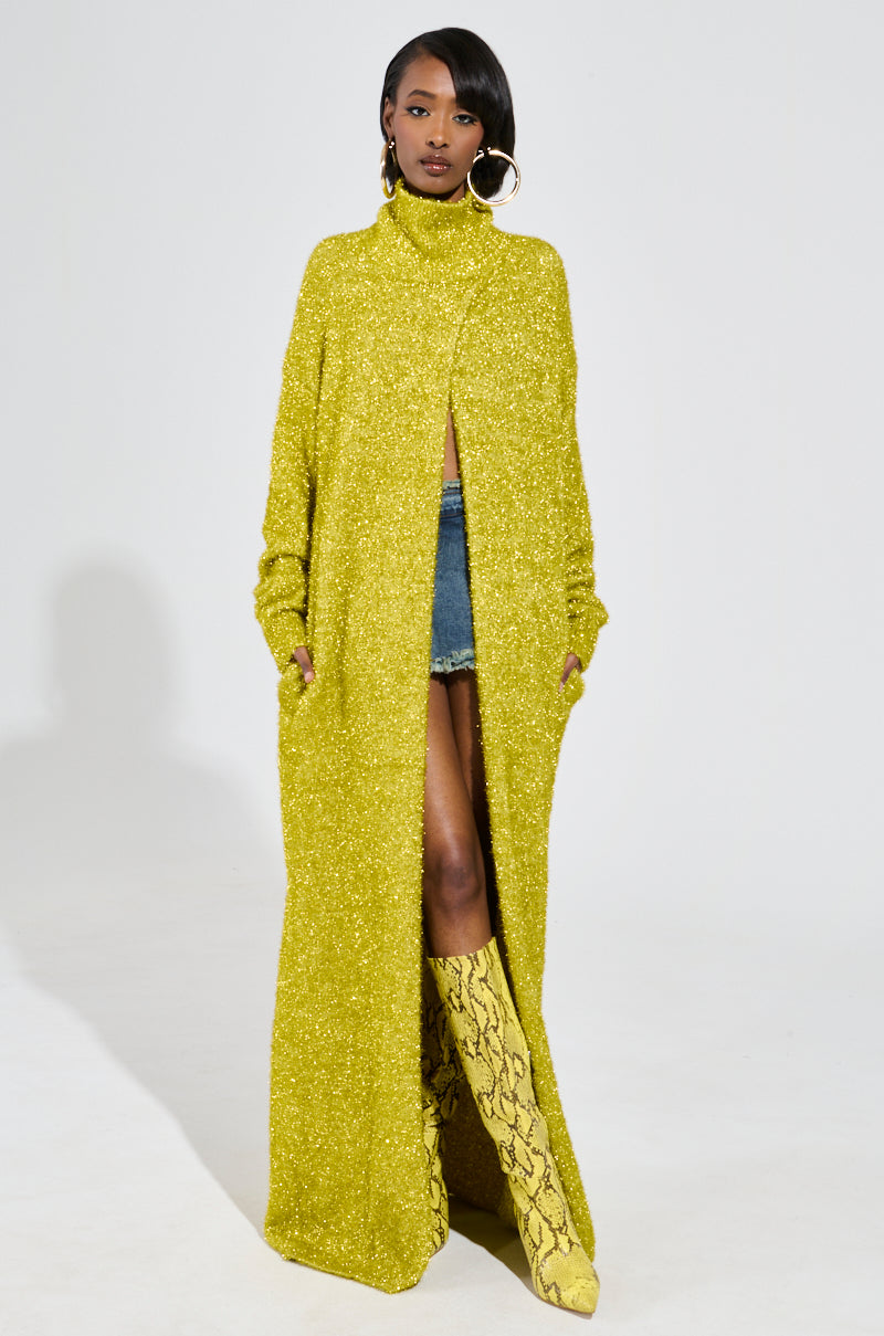 A woman exudes confidence in her MOONLIGHT LONG TURTLENECK CARDIGAN IN GOLD, paired with denim shorts and knee-high yellow snake-patterned boots. Her large hoop earrings gleam as she effortlessly poses against a plain white background.