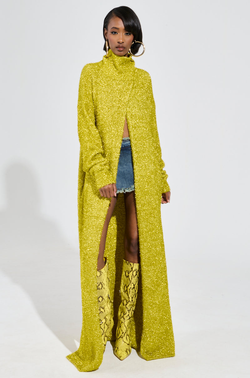 A person models the MOONLIGHT LONG TURTLENECK CARDIGAN IN GOLD, a glittery and stretchy knit design with a high slit that reveals denim shorts. Paired with yellow snake-print boots and large hoop earrings, this maxi-length cardigan stands out against the plain white studio backdrop.