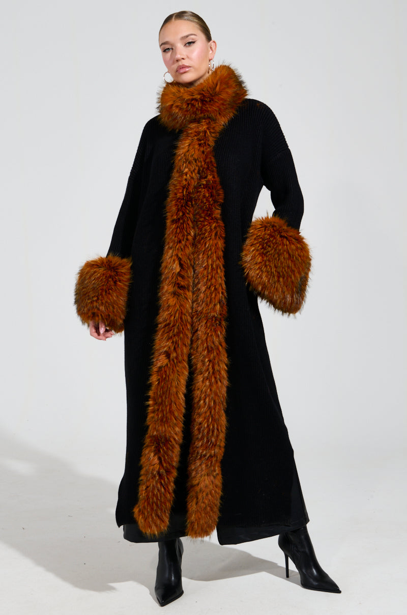 A person wearing the SNOWY NIGHT LONG CARDIGAN IN BLACK, featuring large faux fur accents around the collar and cuffs, stands against a plain white background. With hands resting by their sides and black heeled boots, they perfectly embody a chic winter look.
