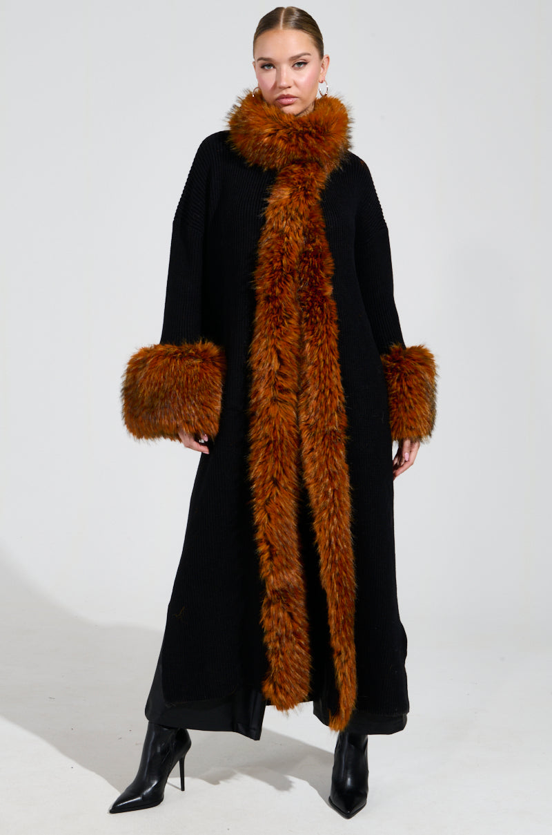 A person wearing the SNOWY NIGHT LONG CARDIGAN IN BLACK, featuring a large, fluffy brown faux fur collar and cuffs, stands against a plain background. They embrace the winter look with black pointed high-heeled boots and have their hair pulled back.
