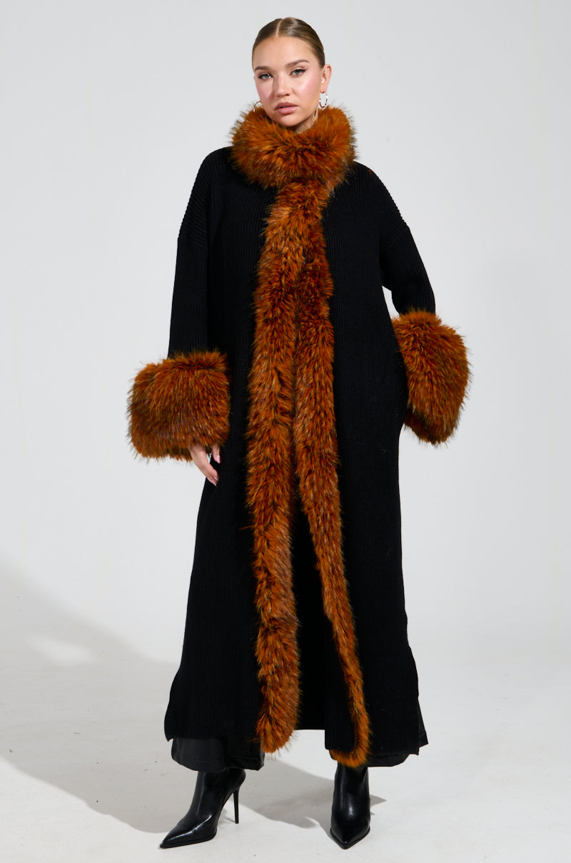 A person showcases a winter look, donning the SNOWY NIGHT LONG CARDIGAN IN BLACK with brown faux fur trim on the collar and sleeves, standing against a plain background. The cardigan features fur detailing down the front and is elegantly paired with black heeled boots.